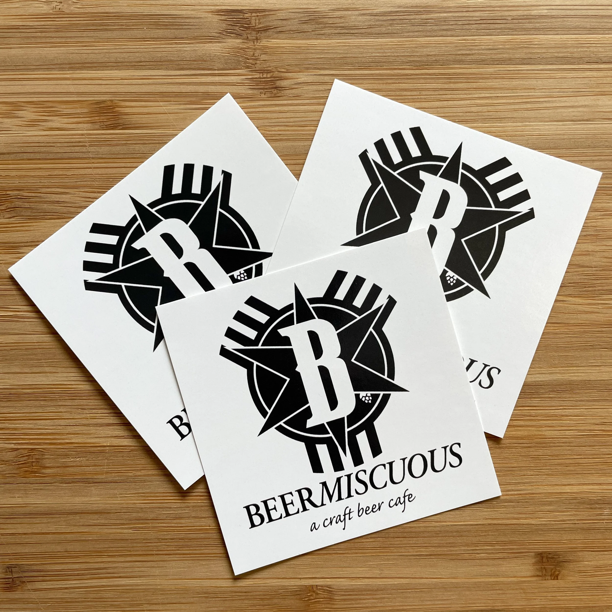 Four white cards with a black compass rose and letter B logo, and text reading "BEER MISC ous" and "a craft beer cafe" on a wooden surface.