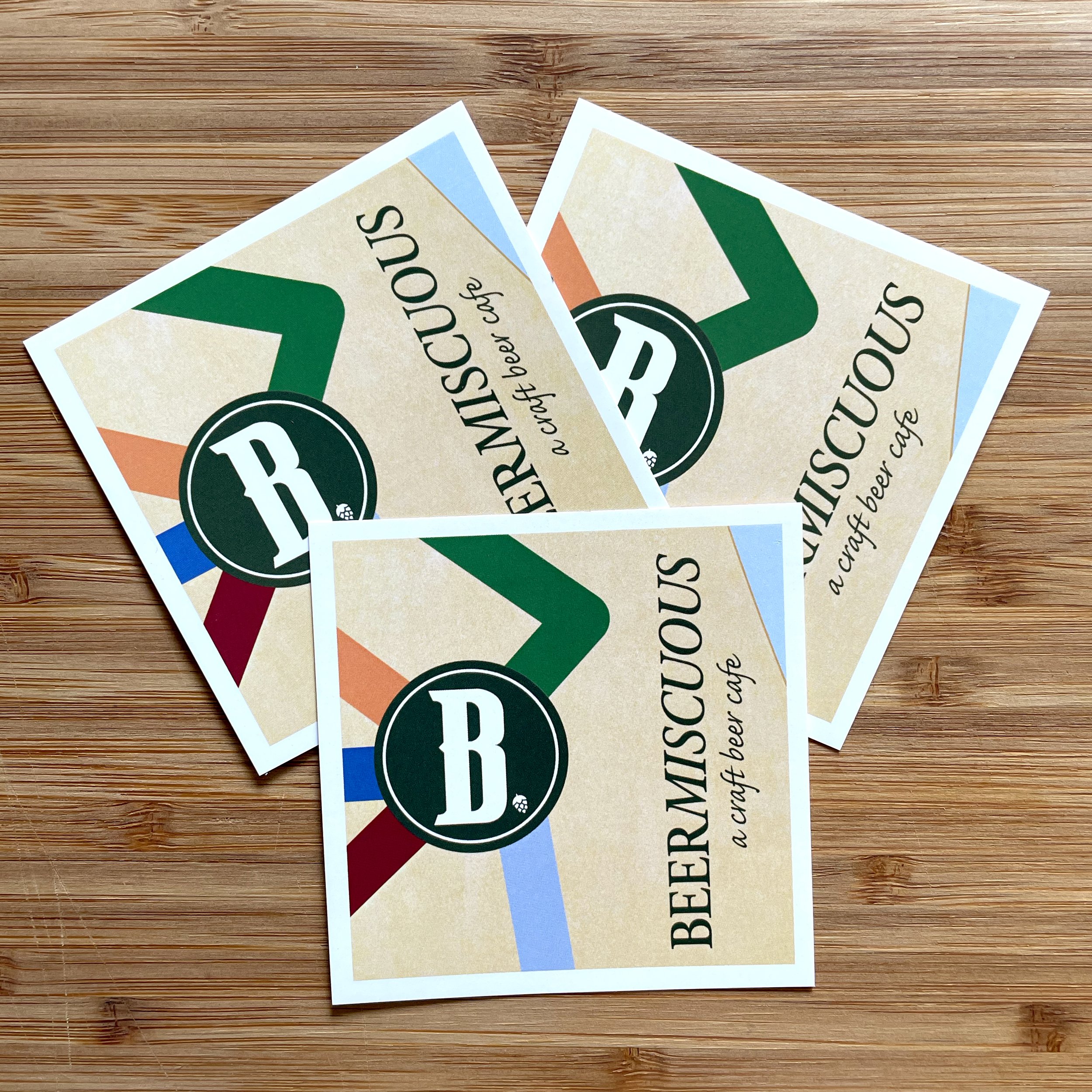 Three beer coasters with a beige background, multicolored stripes, and a black circle with a white letter B in the center, placed on a wooden surface.