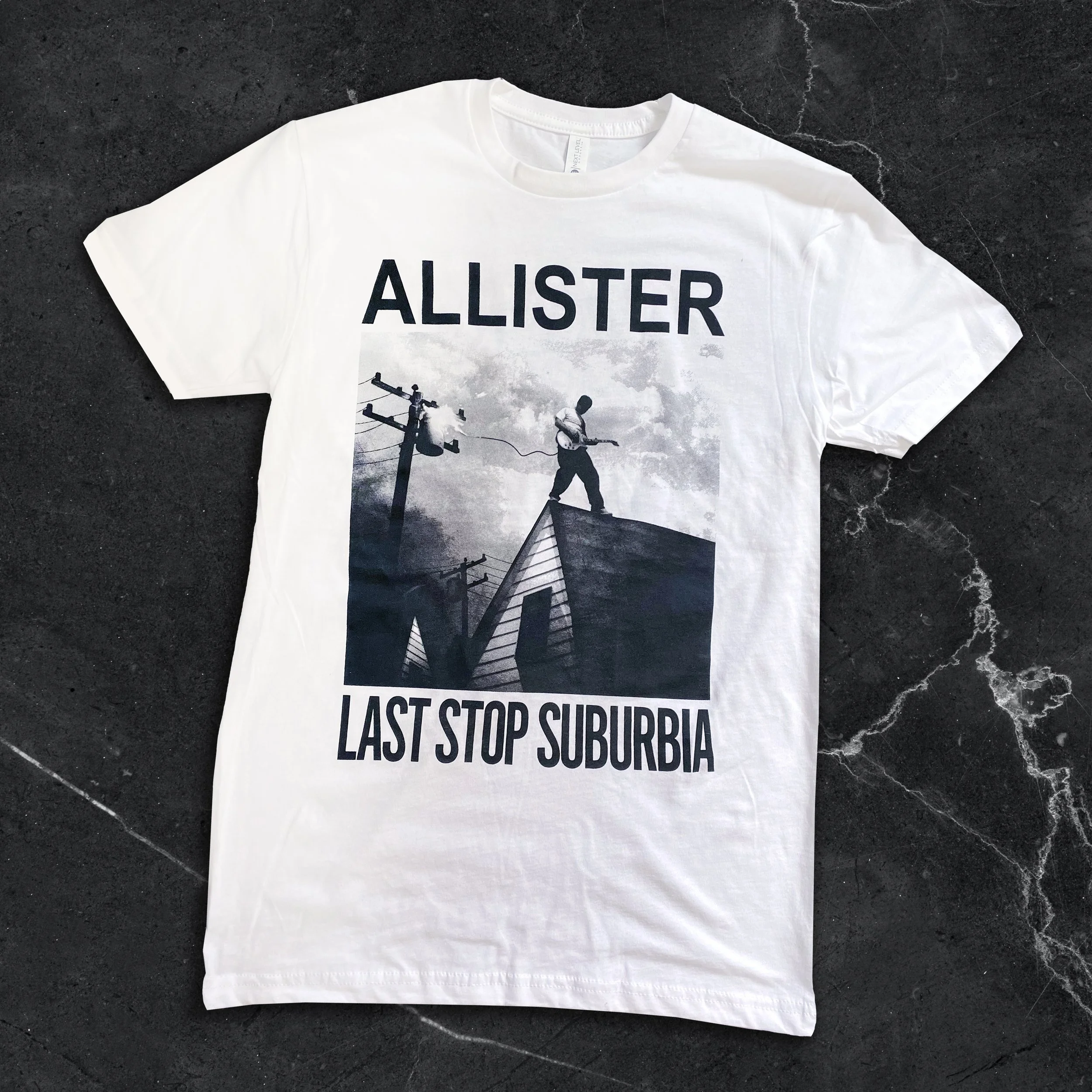 White T-shirt with a black and white graphic of a person standing on a rooftop, holding a guitar, with power lines nearby. The text on the shirt reads 'ALLISTER' at the top and 'LAST STOP SUBURBIA' at the bottom.