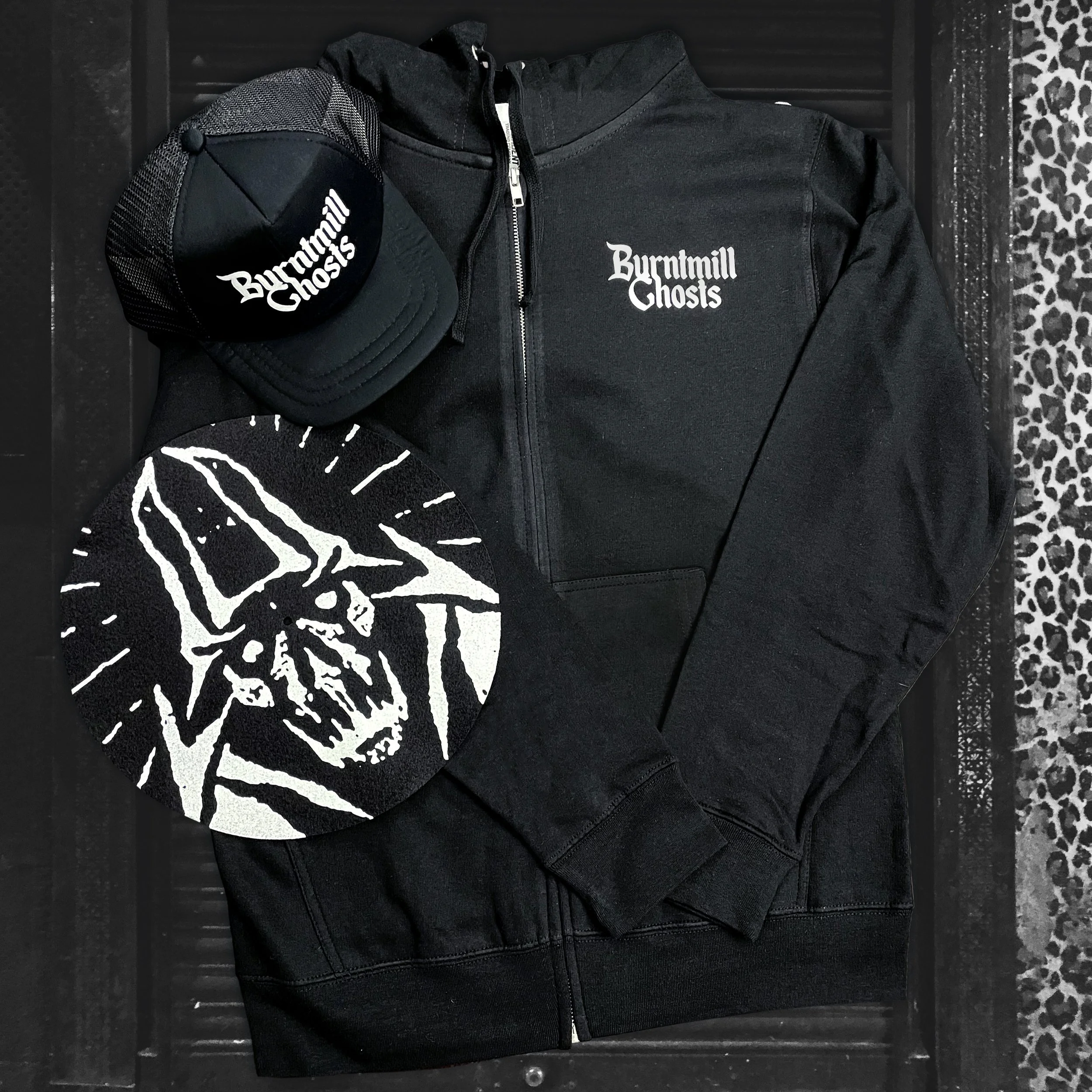Black hoodie with 'Burntmill Ghosts' embroidered on the chest, a black mesh-back trucker cap with 'Burntmill Ghosts' written on the front, and a round patch featuring a stylized skull design, all displayed on a dark background with a leopard print bo