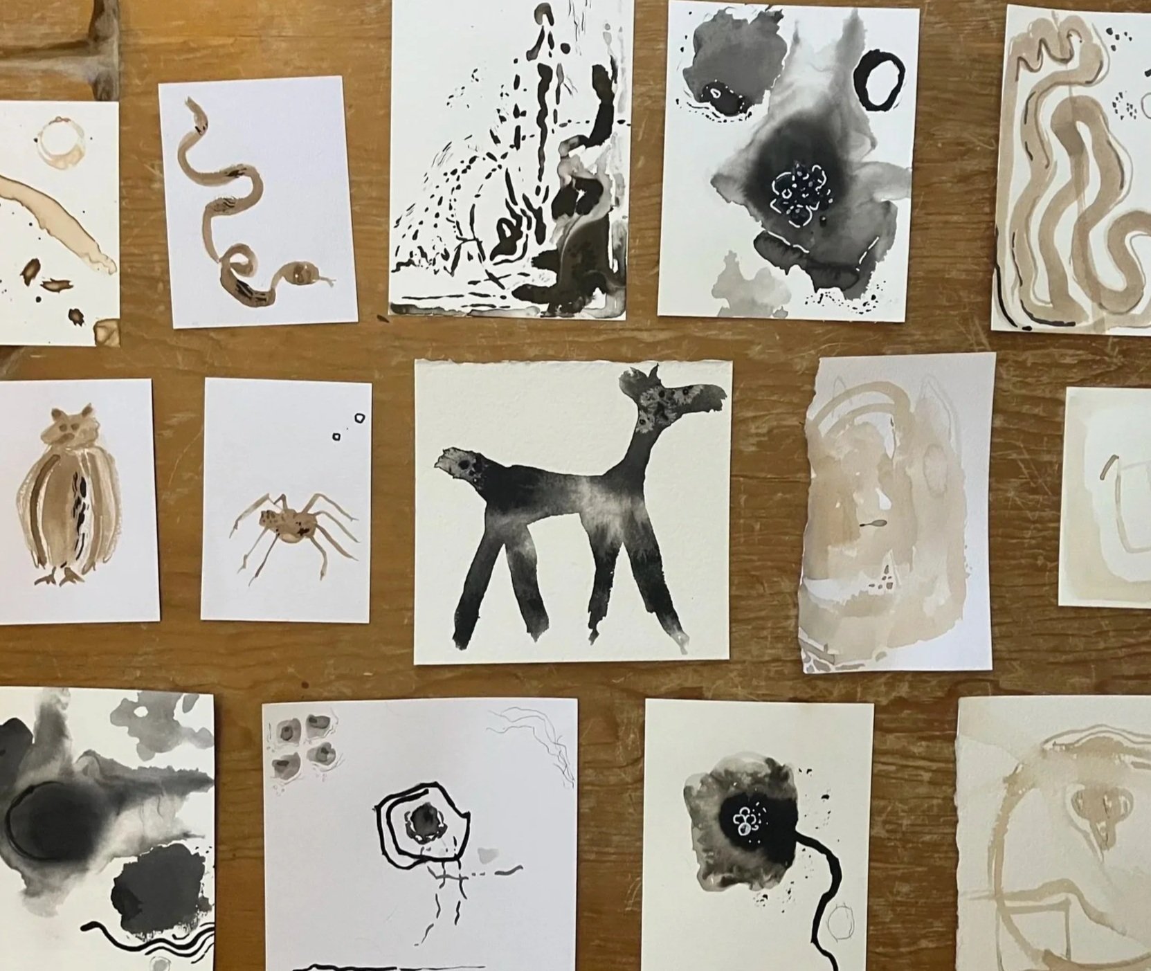Collection of ink paintings on small paper sheets depicting animals and abstract designs, including a fox, a snake, a spider, and a bird, arranged on a wooden surface.