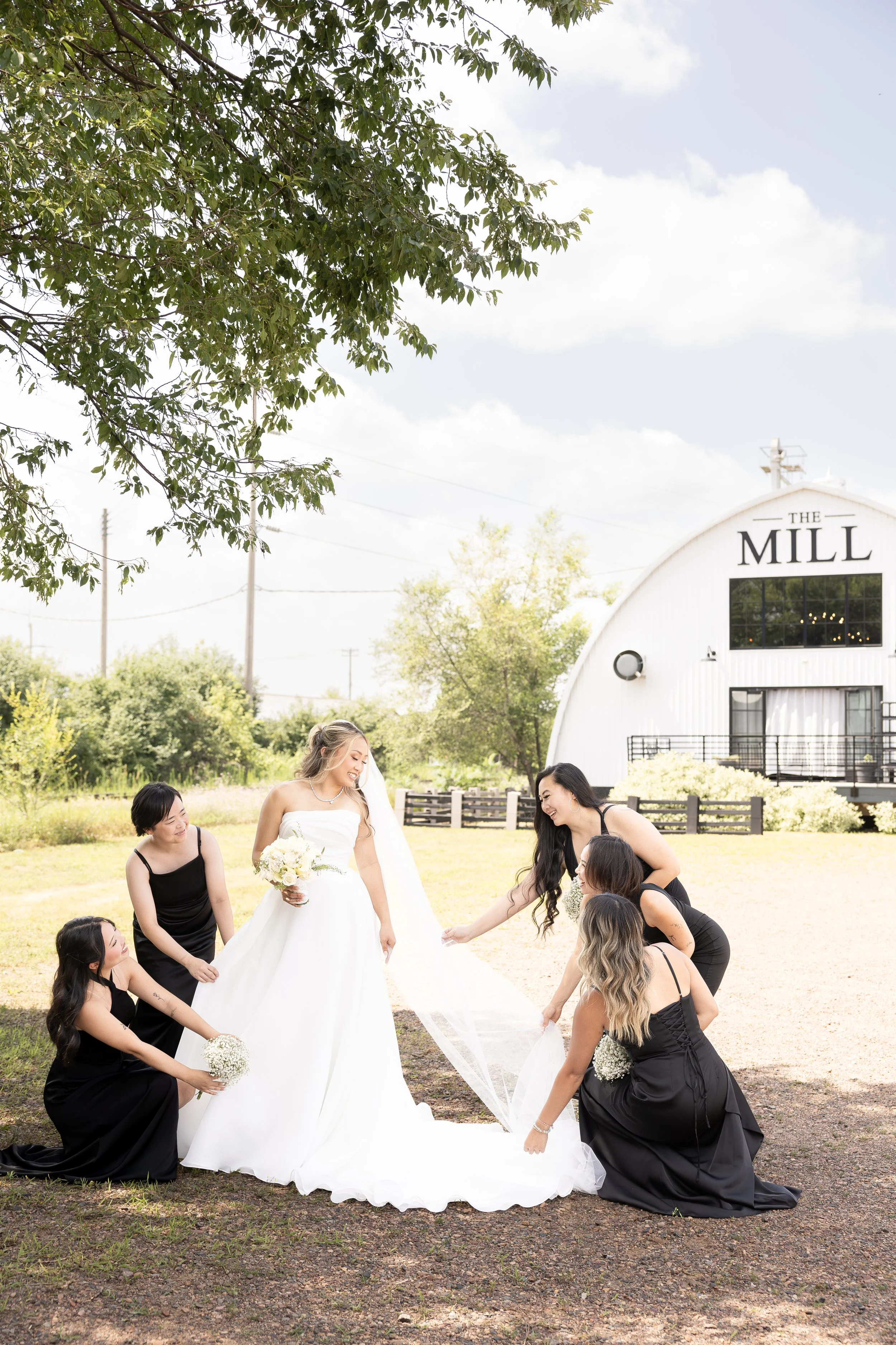 A bridal party outside with a bride in a white wedding gown holding a bouquet, surrounded by women in black dresses, some holding white flowers, as they lift and arrange her train on a grassy area near a white building labeled 'The Mill' under a part