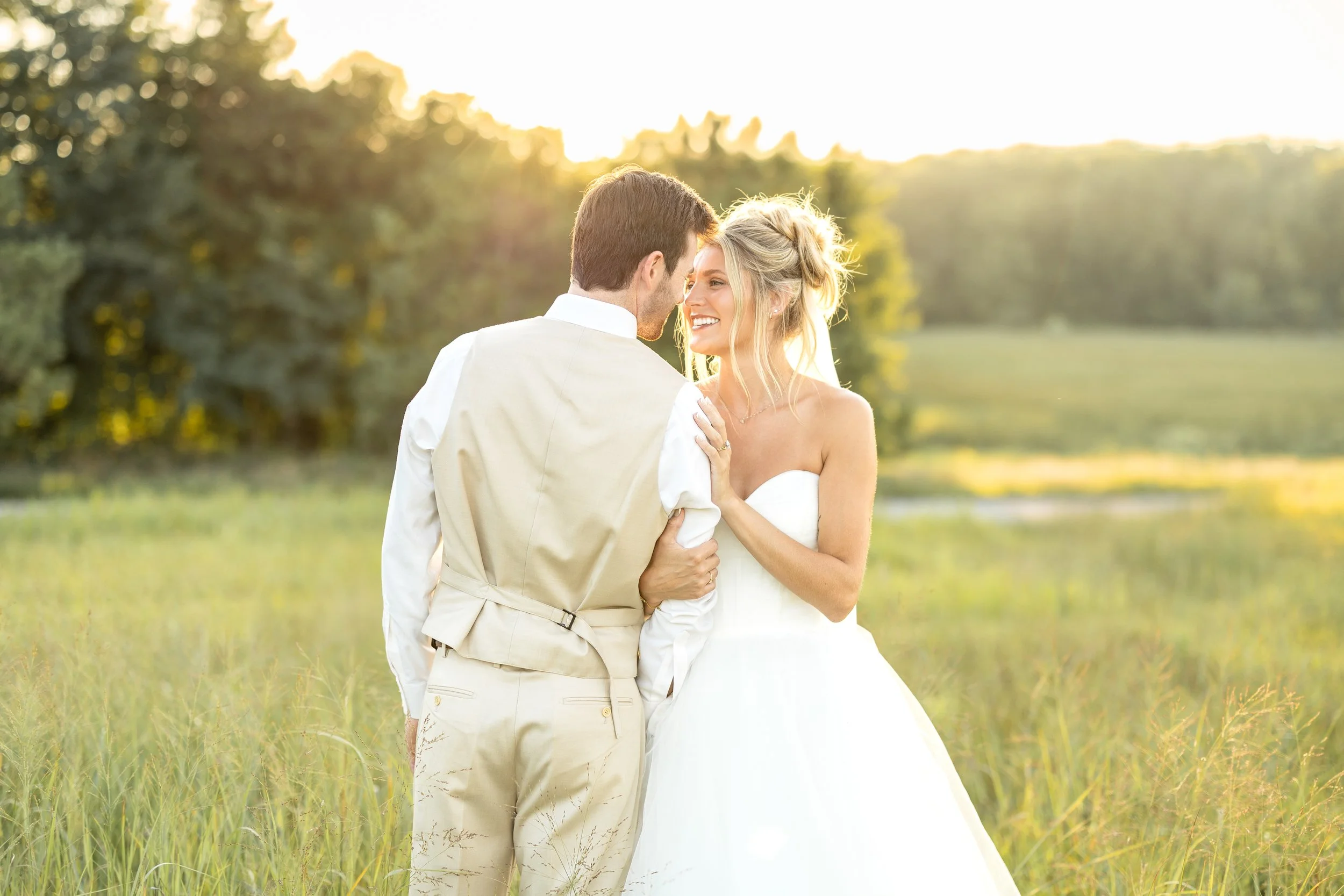 A bride and groom standing close together outdoors during sunset, embracing each other and smiling affectionately, with lush greenery and trees in the background.