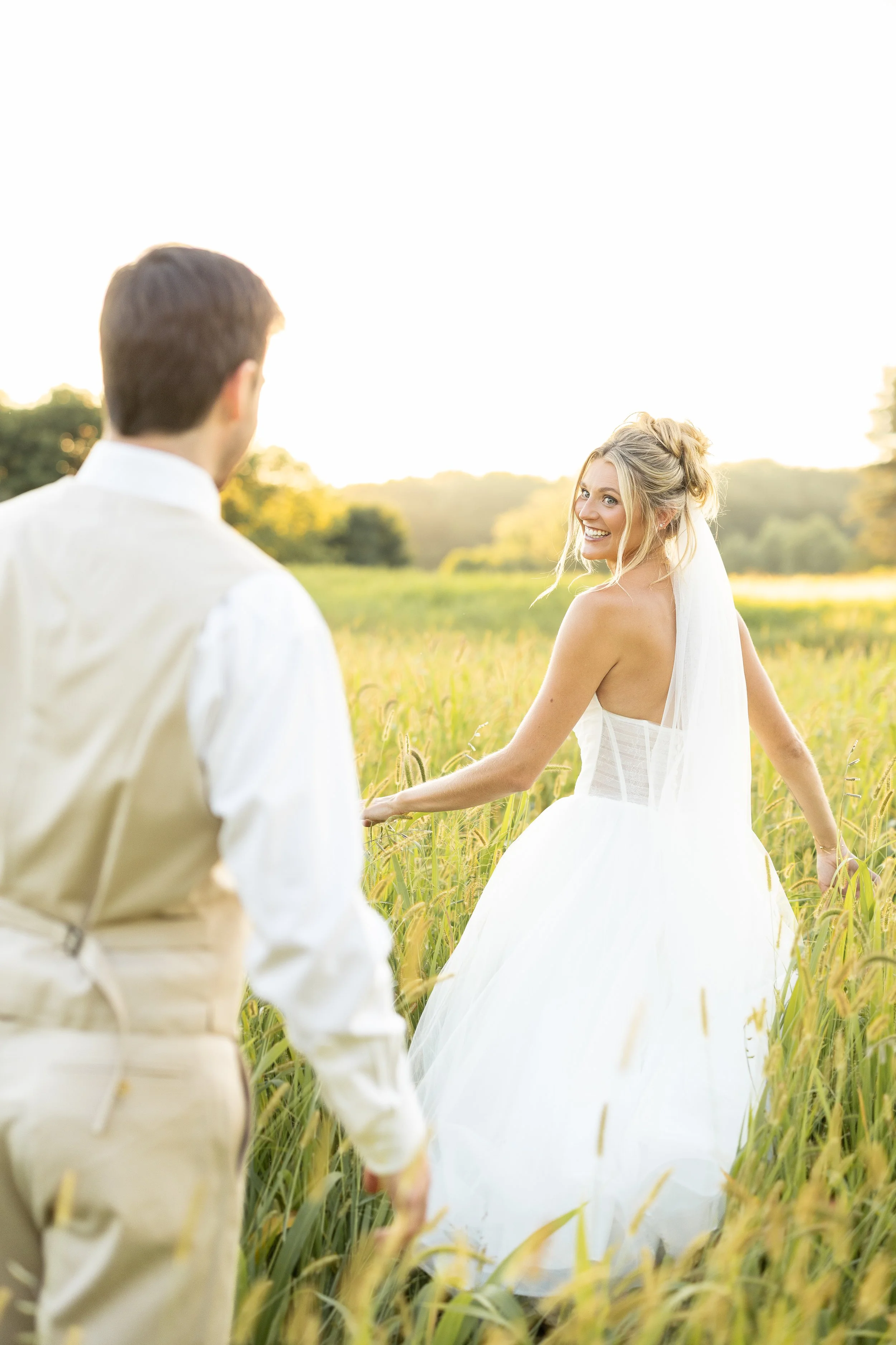 A bride smiling at a groom in a field, holding hands, with the sun setting in the background.