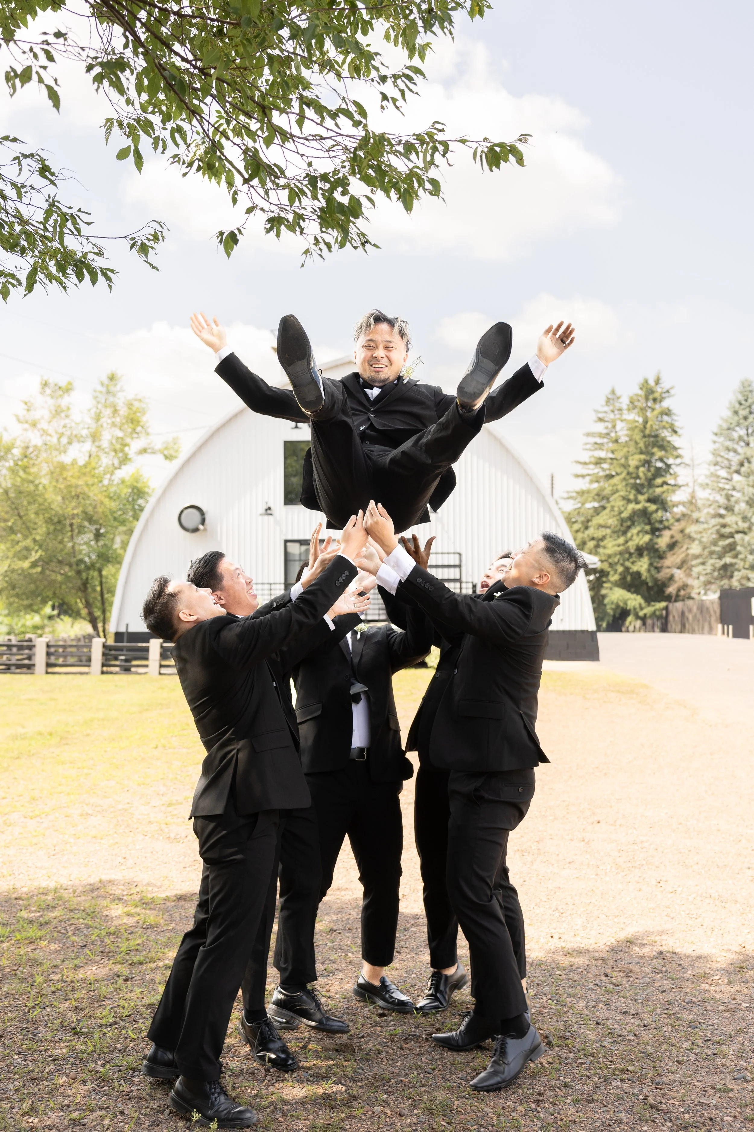 Group of people dressed in black suits lifting and tossing a man in a wedding tuxedo with a joyful expression during an outdoor celebration.