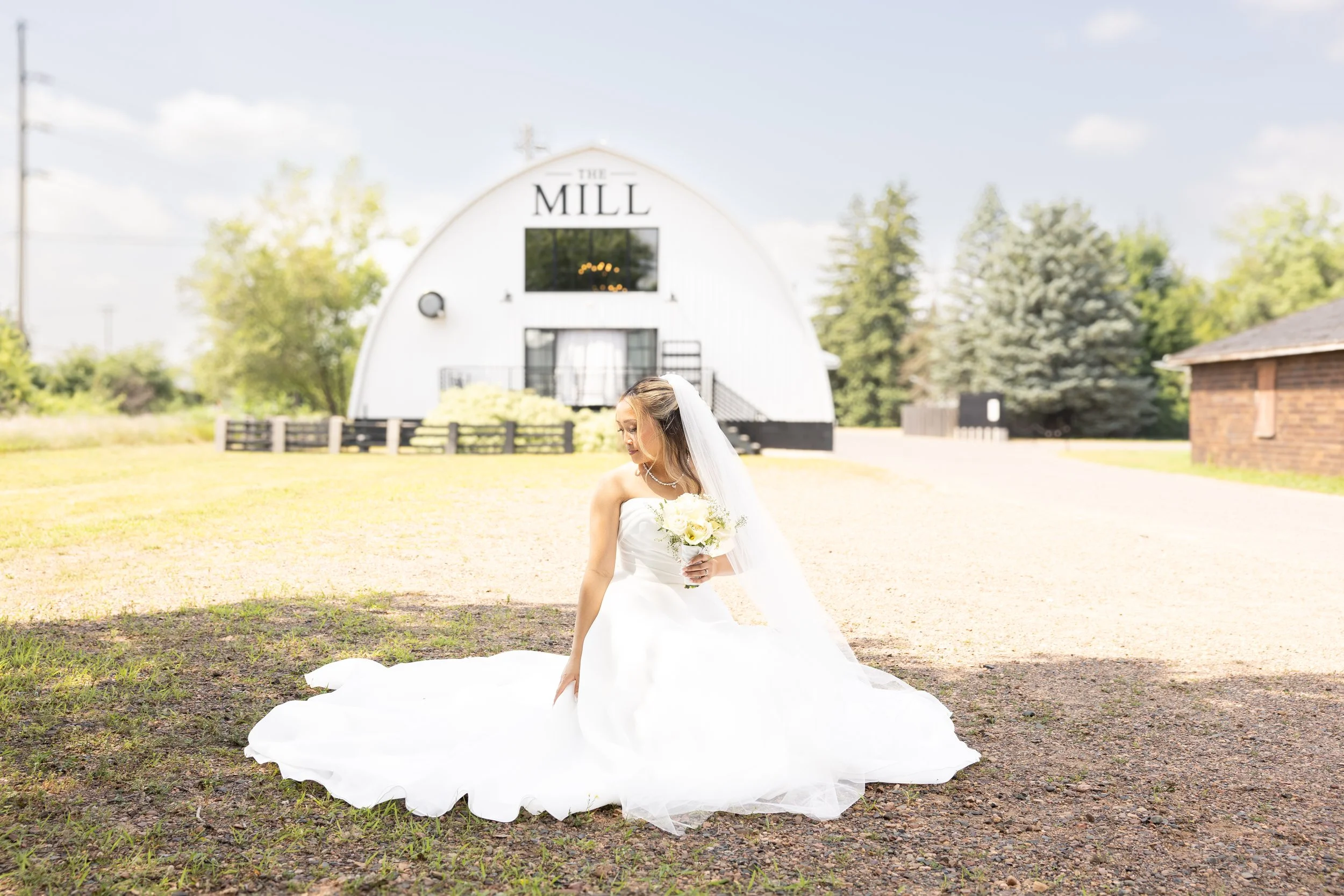 A bride in a white wedding gown holding a bouquet of white roses, standing outdoors on a sunny day with a white barn labeled "The Mill" in the background, surrounded by trees.