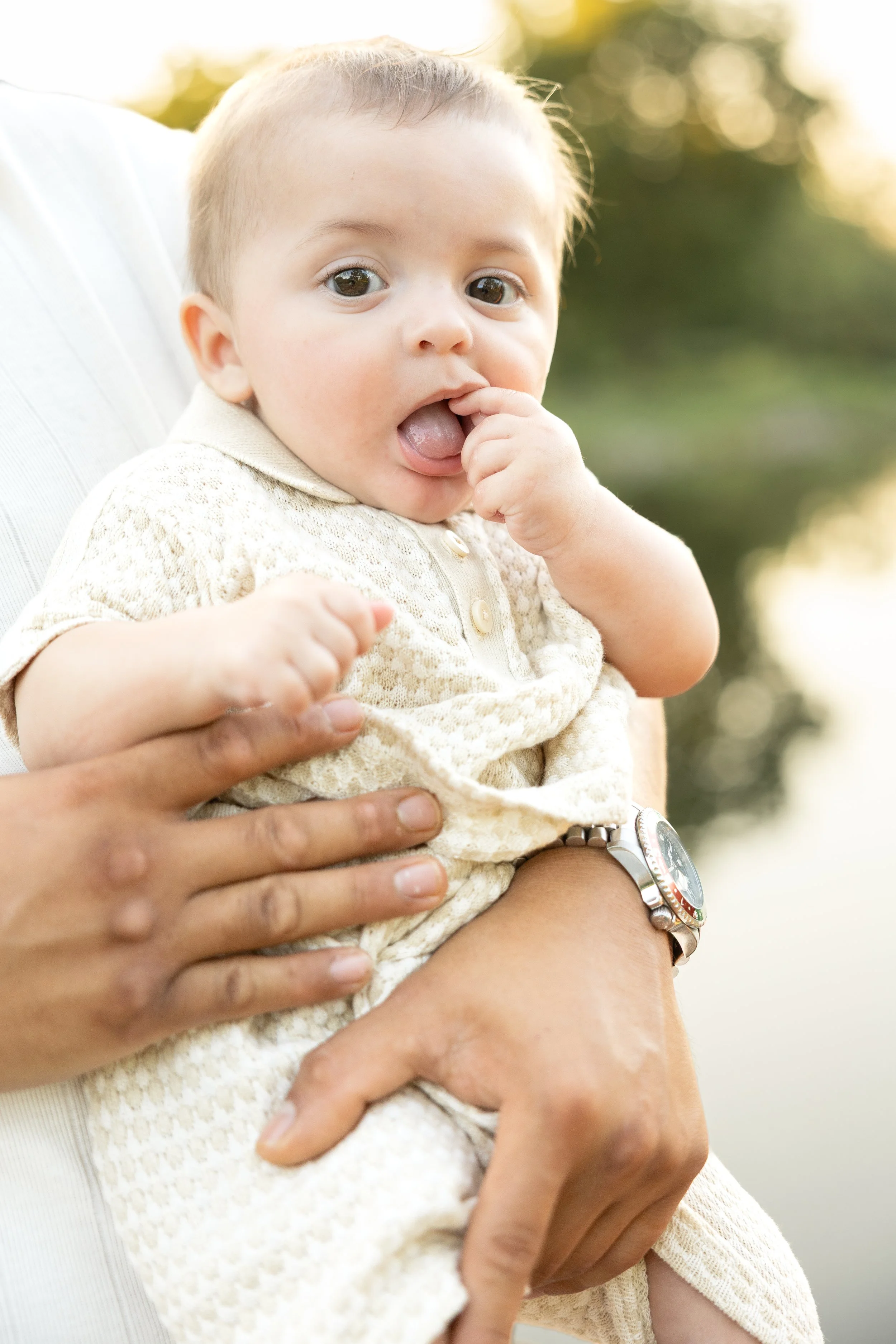 A close-up of a smiling baby with big eyes, wearing a light-colored, textured shirt, with someone holding him, outdoors in natural light.