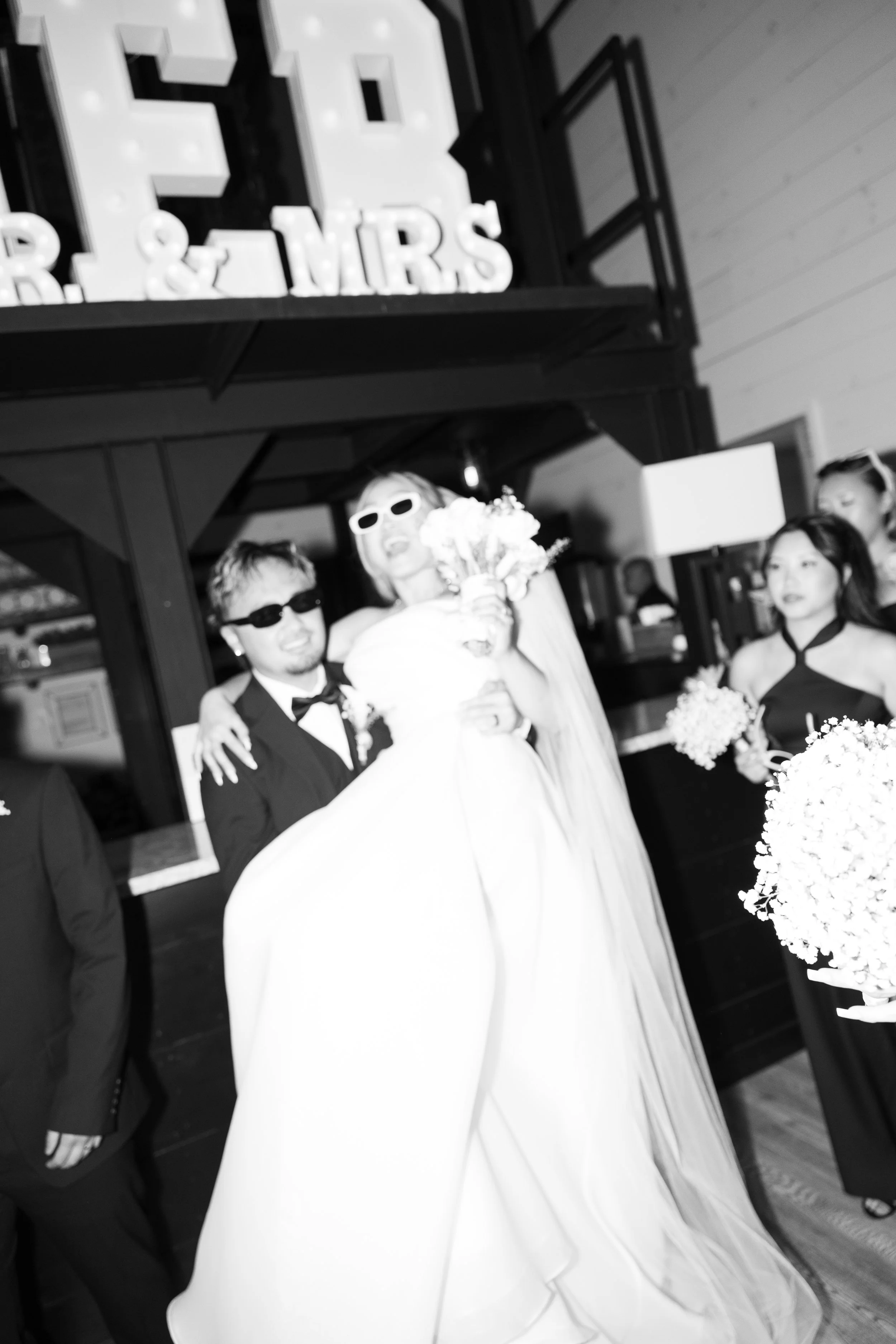 A couple wearing sunglasses, with the woman dressed in a wedding gown and the man in a tuxedo, at a wedding reception with decorative signage in the background.
