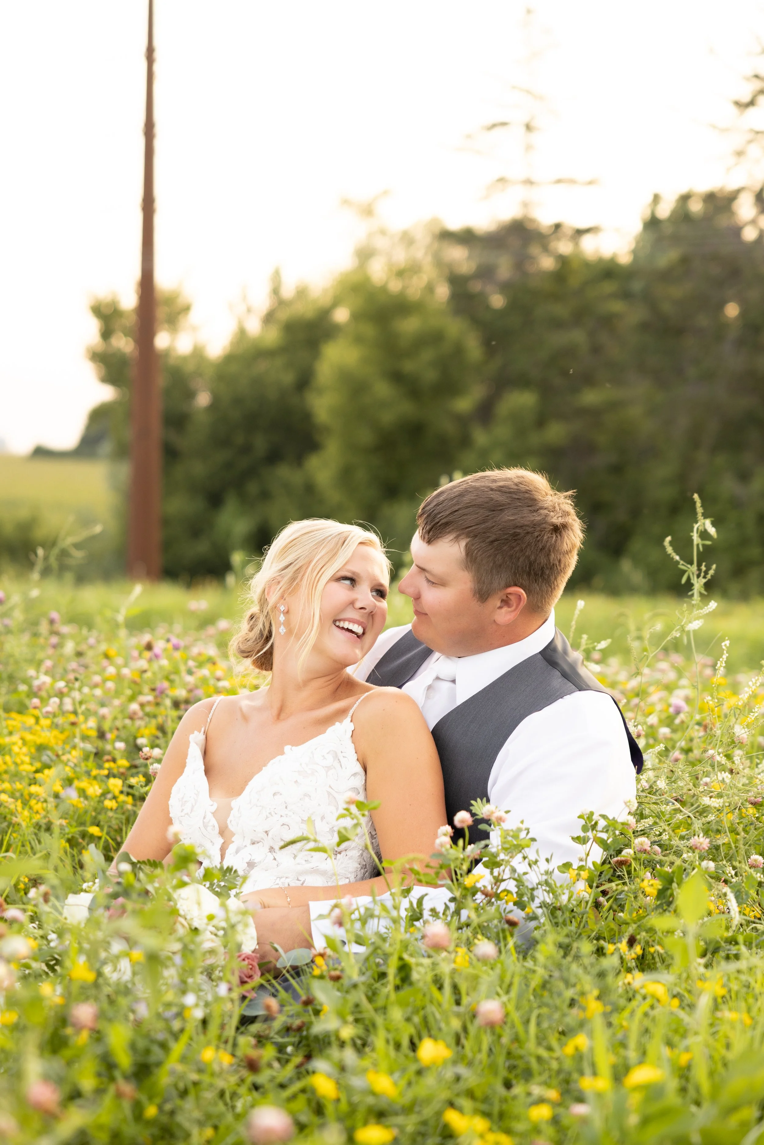 A bride and groom sitting in a field of colorful wildflowers, smiling and looking into each other's eyes during sunset, with trees and a utility pole in the background.