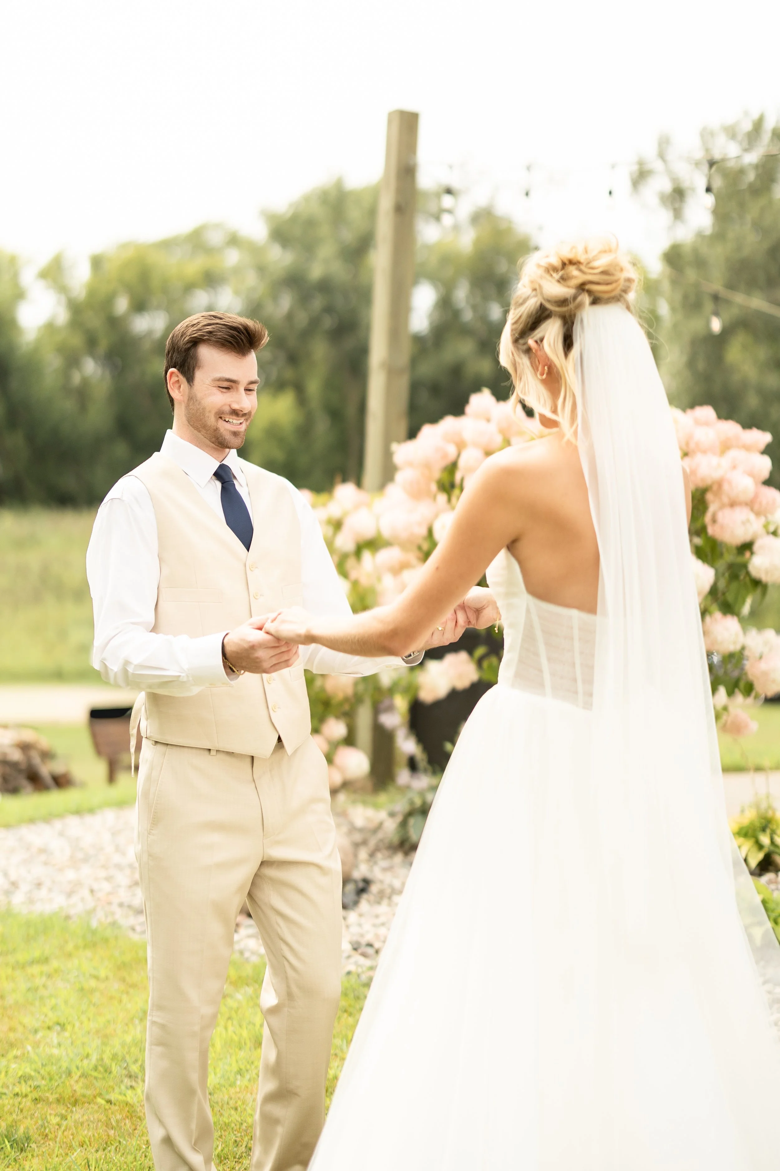 A couple exchanging wedding vows outdoors, with the groom smiling and holding the bride's hands, surrounded by pink flowers and greenery.