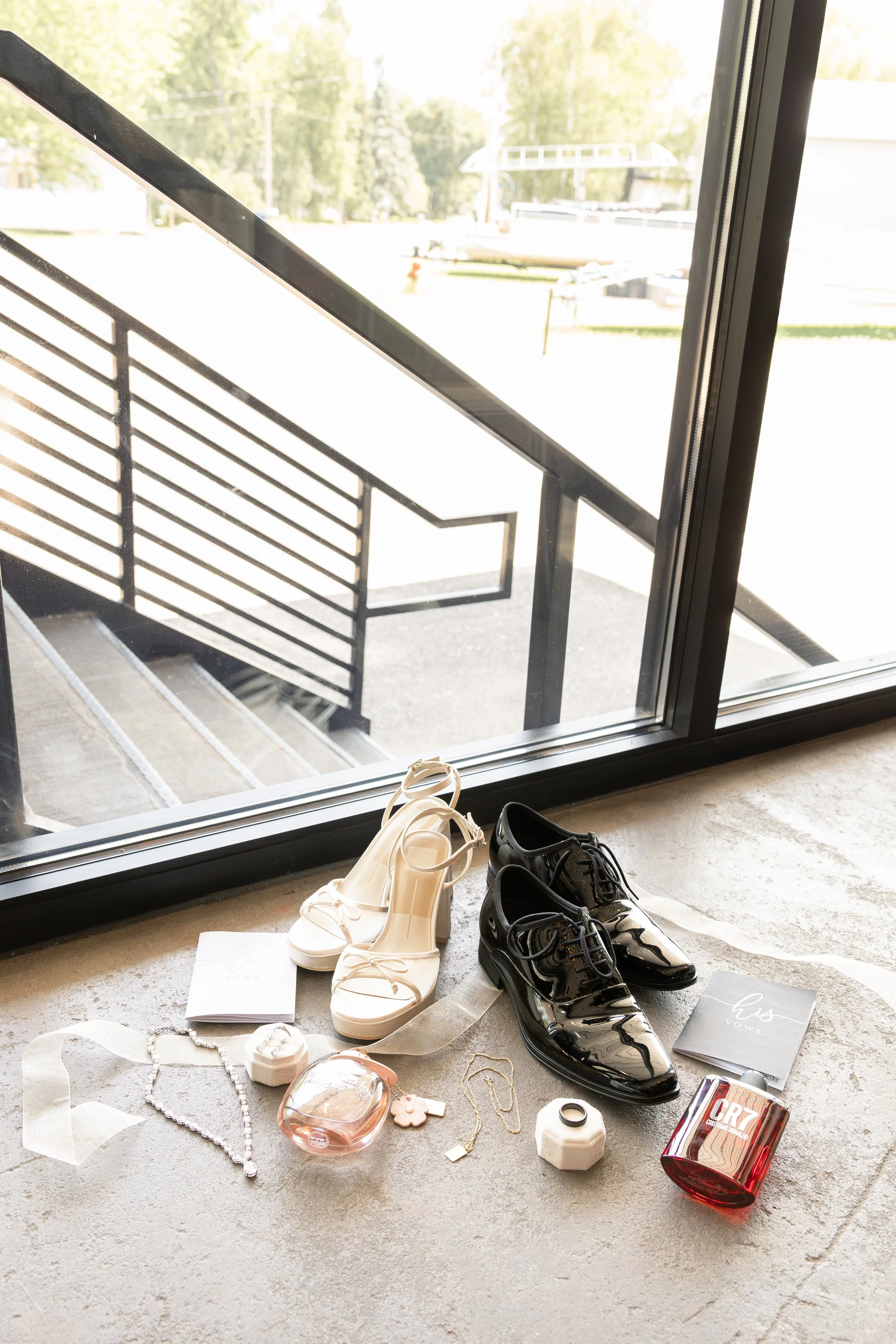Shoes, jewelry, perfume, and other accessories displayed on the floor near a glass door leading to an outdoor balcony with a metal railing, seen in daylight.