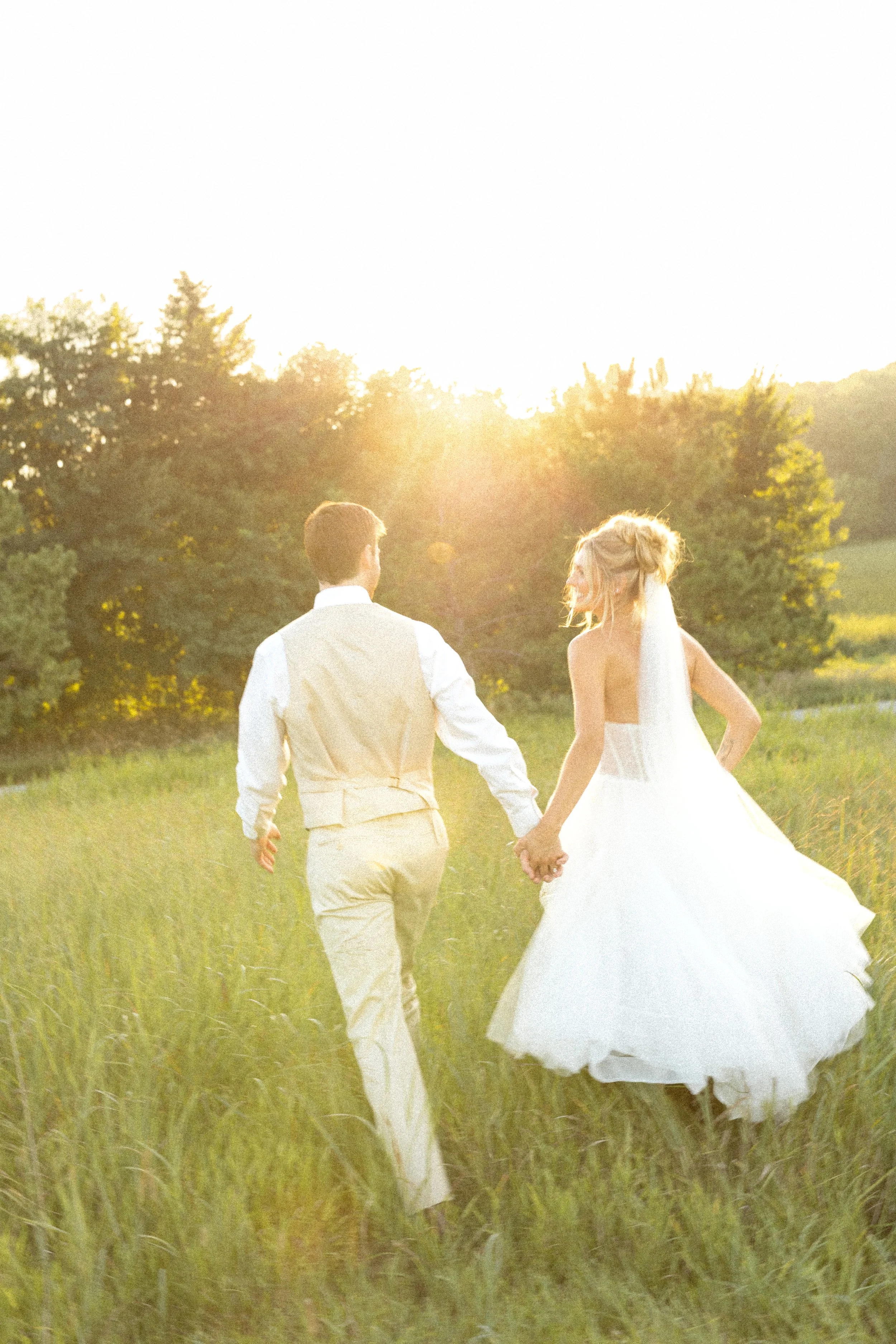 A bride and groom holding hands and walking through a grassy field during sunset.