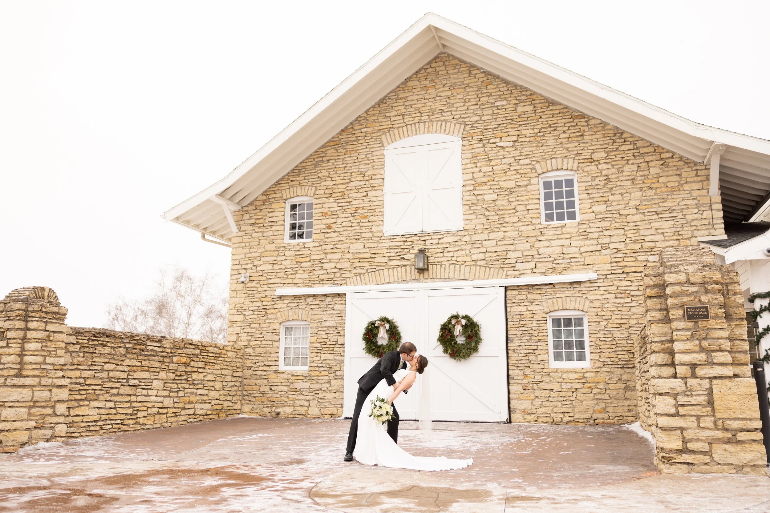 A bride and groom sharing a kiss in front of a rustic stone barn decorated for Christmas with wreaths.