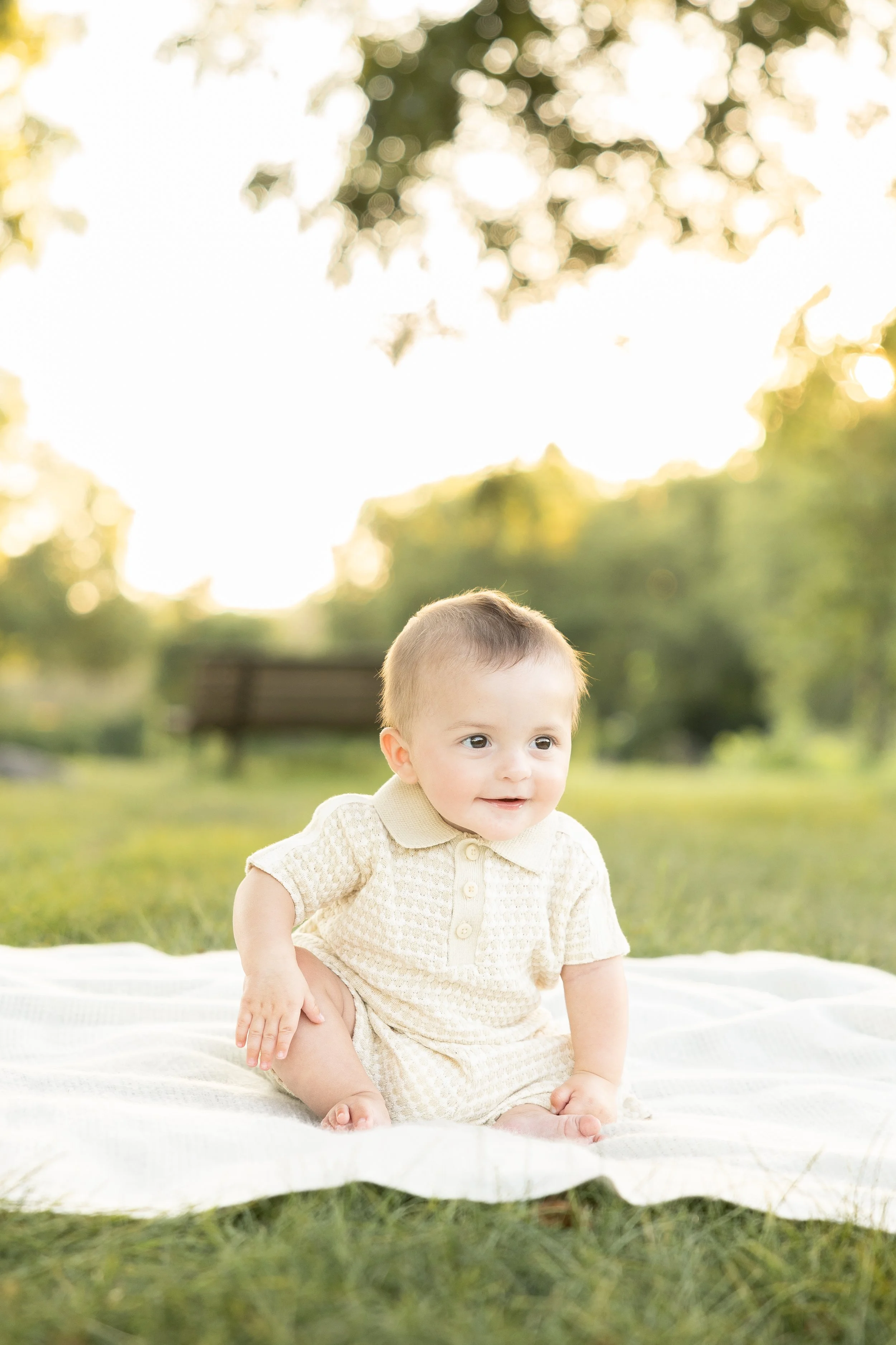 A young child with short light brown hair sitting on a white blanket outdoors in a park, with green grass and trees, during sunny weather.