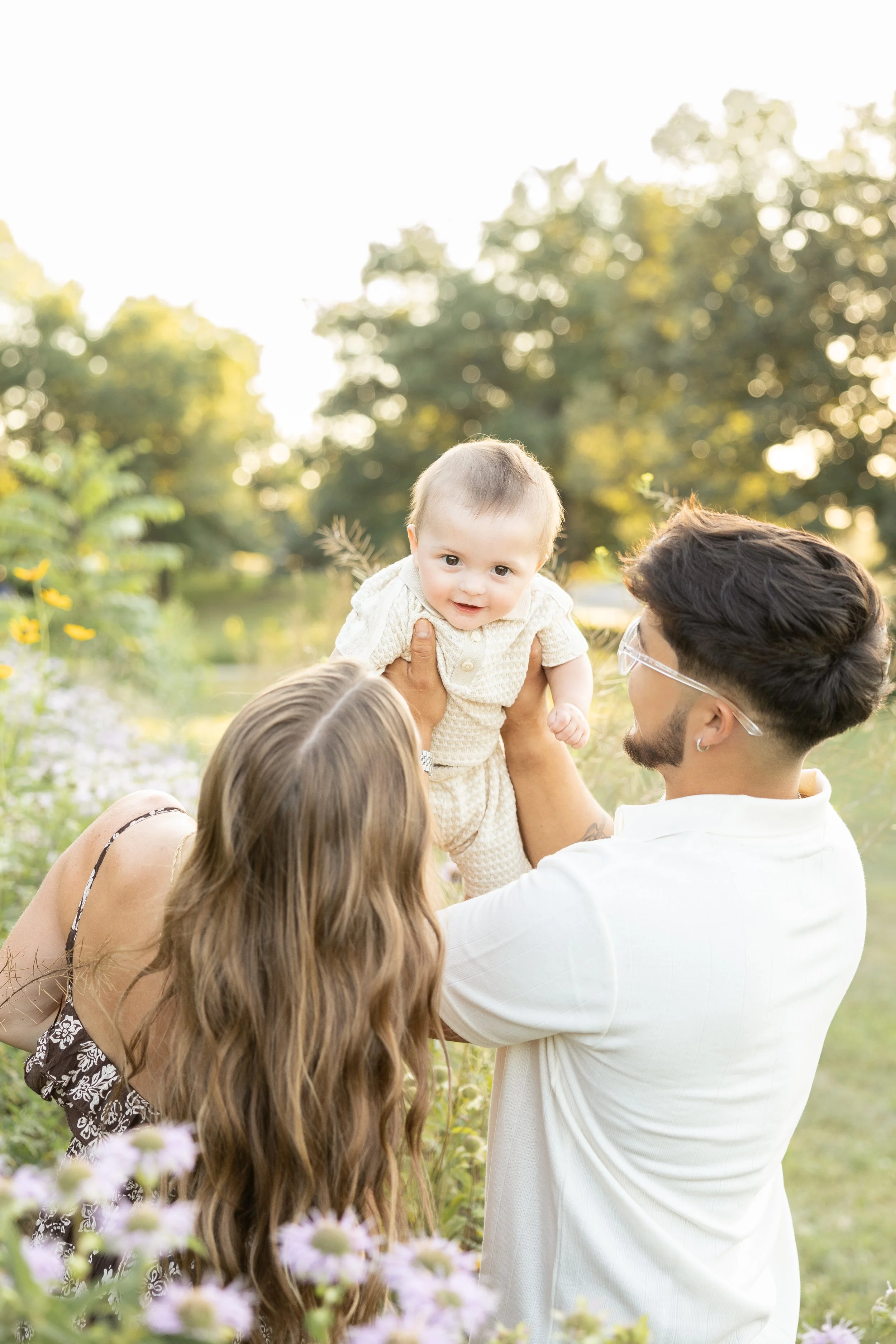 A man lifting a baby in an outdoor setting with trees and flowers in the background during sunset, with two women watching.