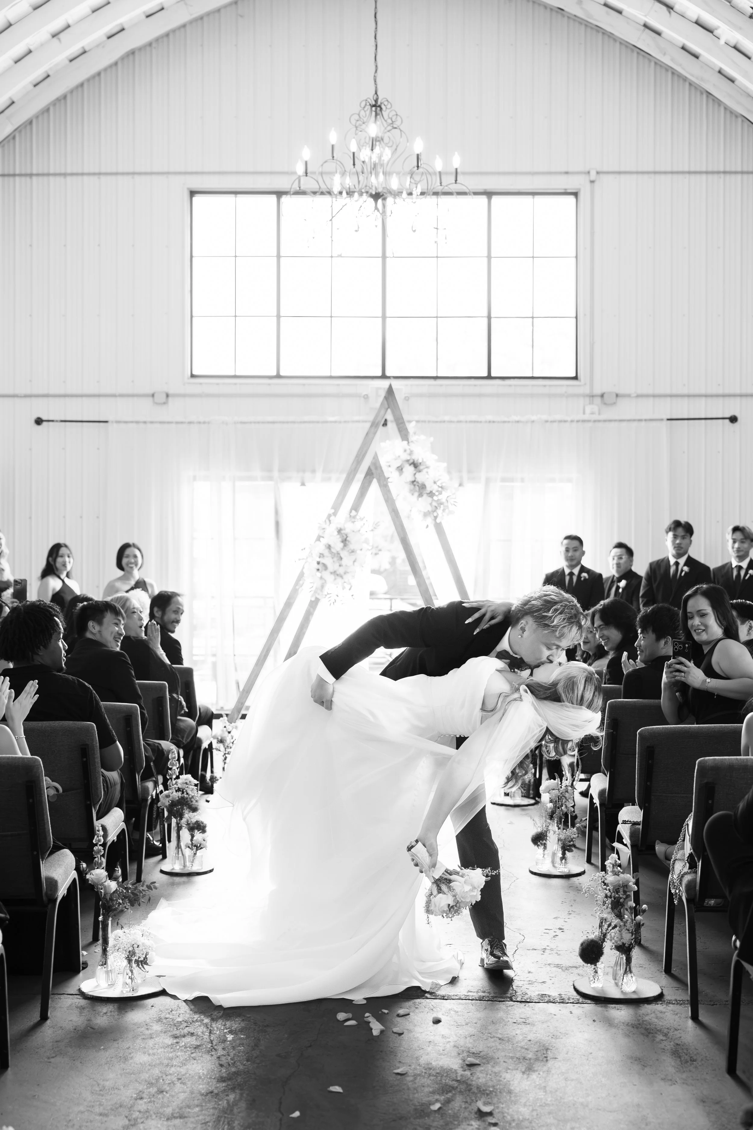 A wedding ceremony with the bride and groom kissing during the vows, surrounded by seated guests in a bright, spacious hall with large windows and a chandelier.