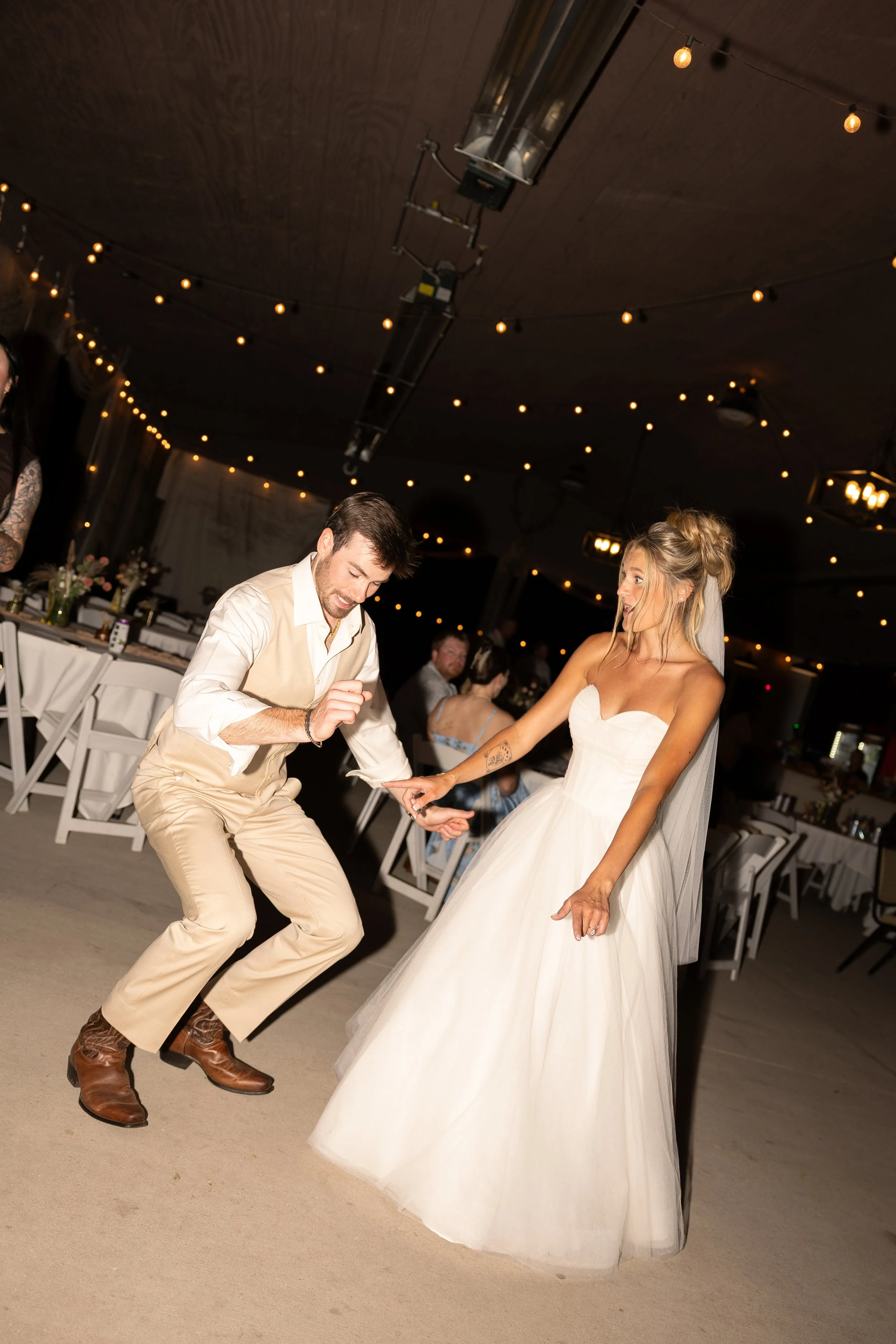 A bride and groom dancing at a wedding reception with string lights overhead and tables set for guests in the background.