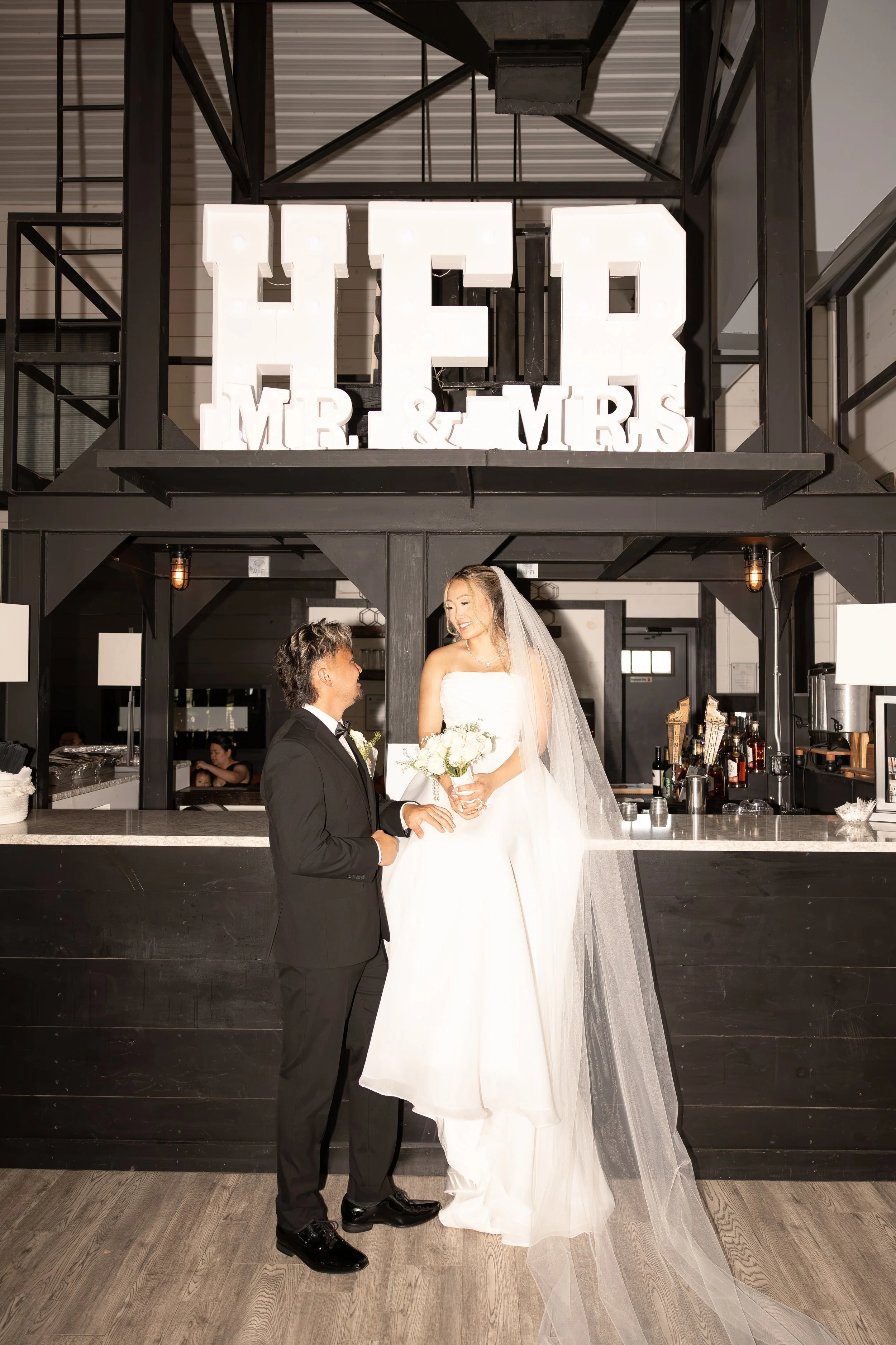 A bride and groom are standing in front of a bar, smiling at each other. The bride is wearing a white wedding gown with a long veil and holding a bouquet of white flowers. The groom is dressed in a black tuxedo with a bow tie. Behind them, large deco