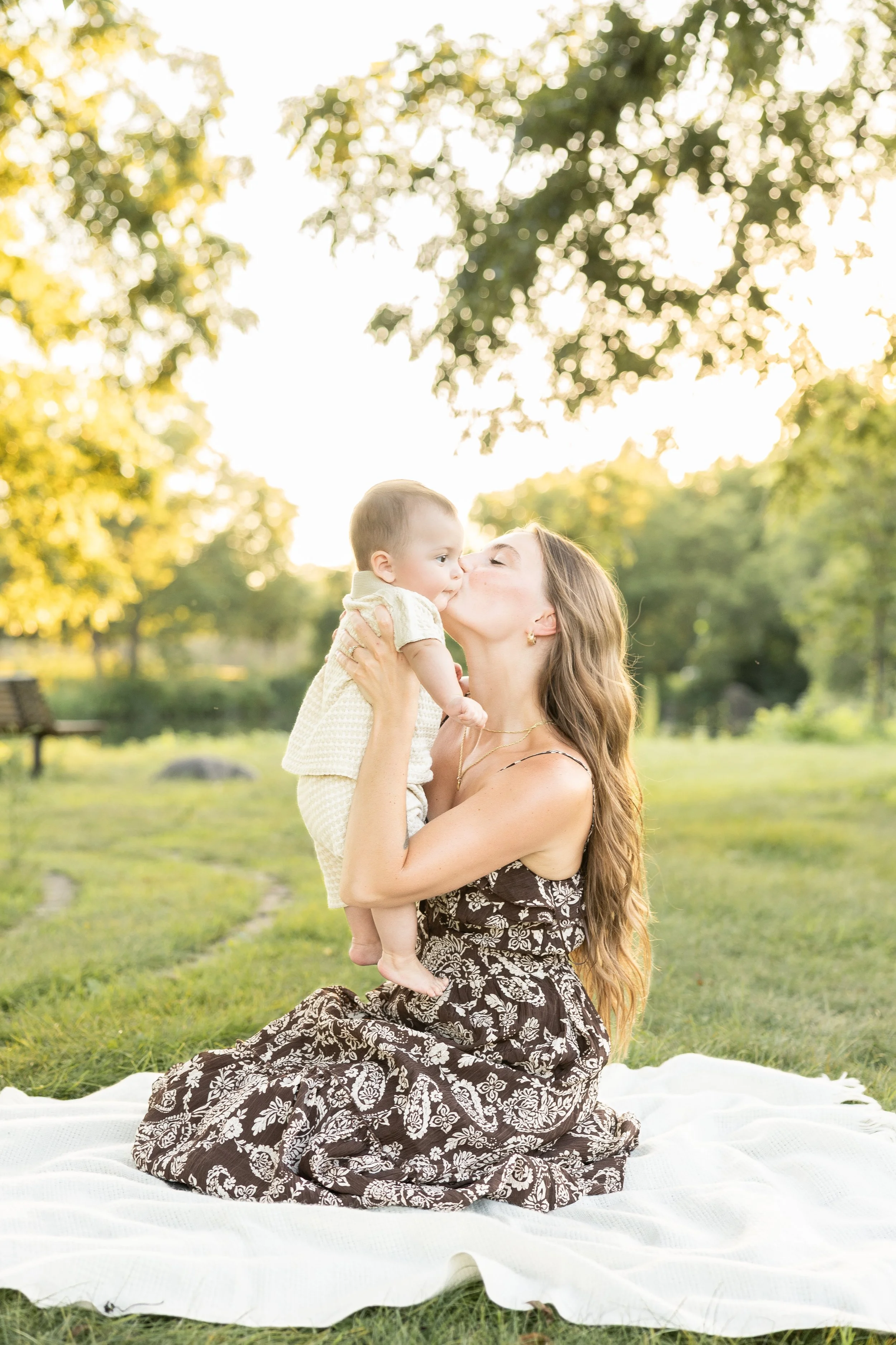 A woman with long hair, wearing a black and white floral dress, kneels on a white blanket in a park, holding a young child close to her face for a kiss, with green trees and sunlight in the background.