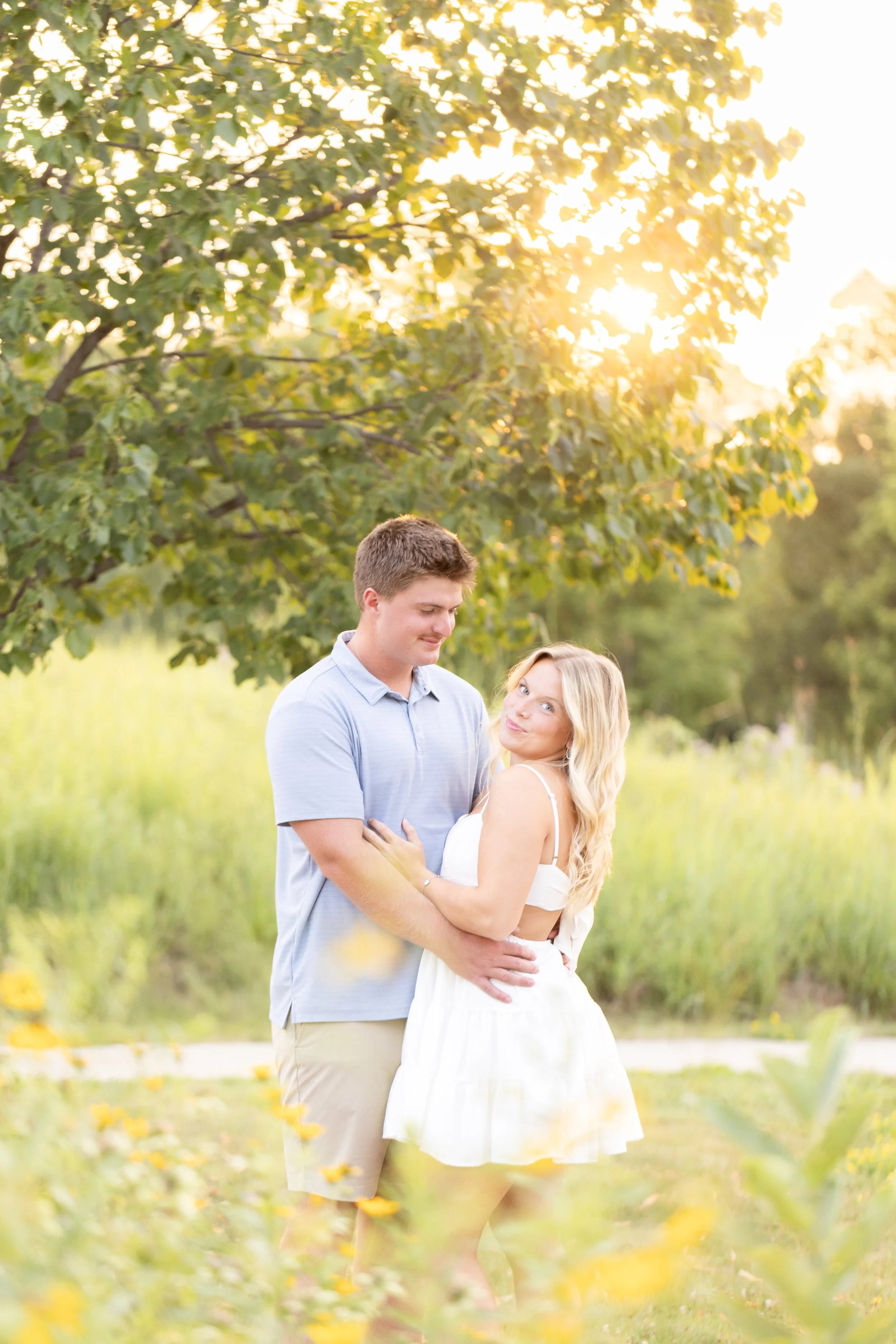 A young couple stands closely together in a grassy outdoor setting during sunset, with green trees and yellow flower petals scattered on the ground surrounding them.