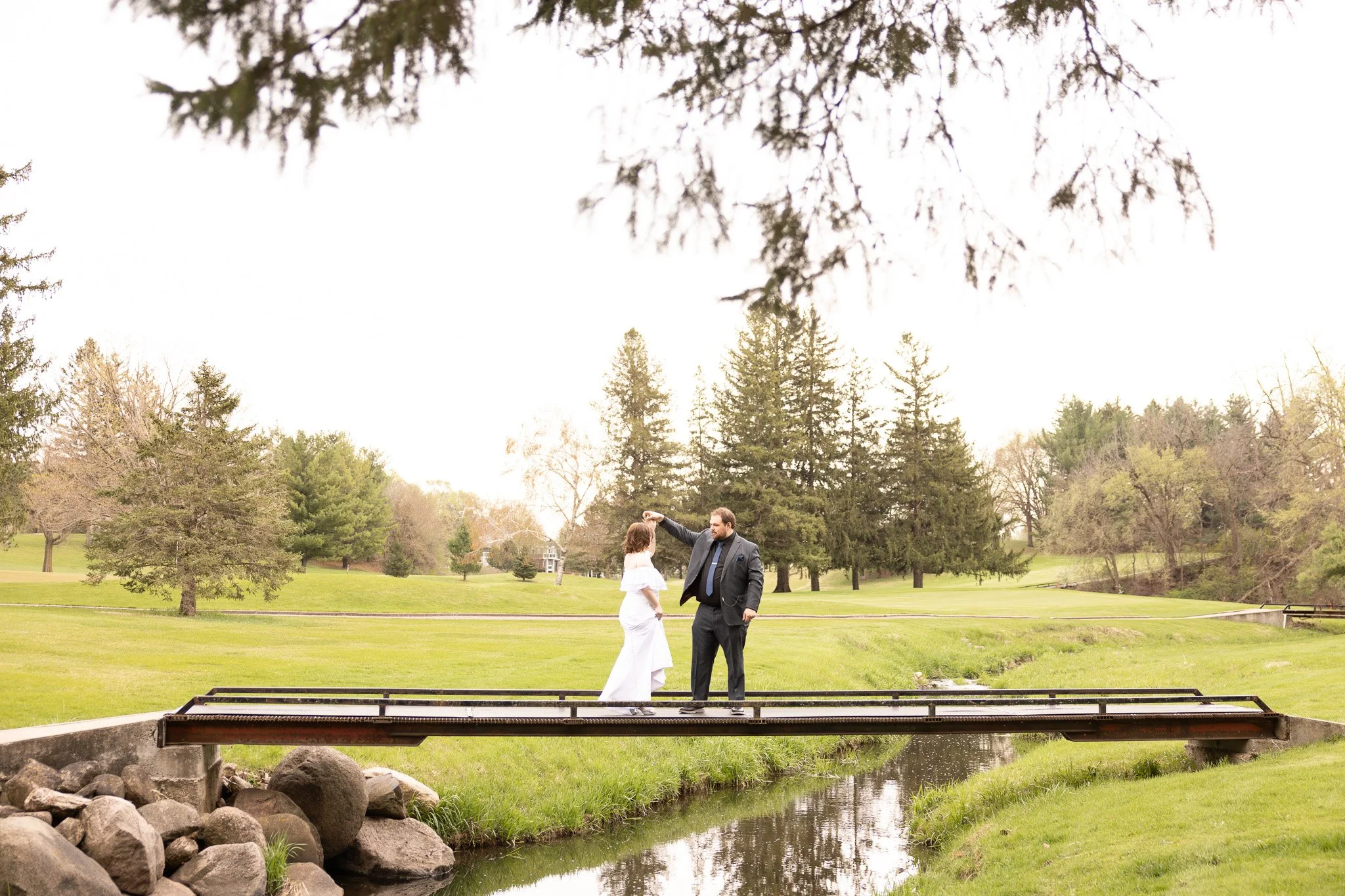 A couple are getting married outdoors in a park. The bride is in a white dress and the groom is in a dark suit. They are holding hands under a large tree and the groom is touching the bride's head. There is a small stream with rocks in the foreground