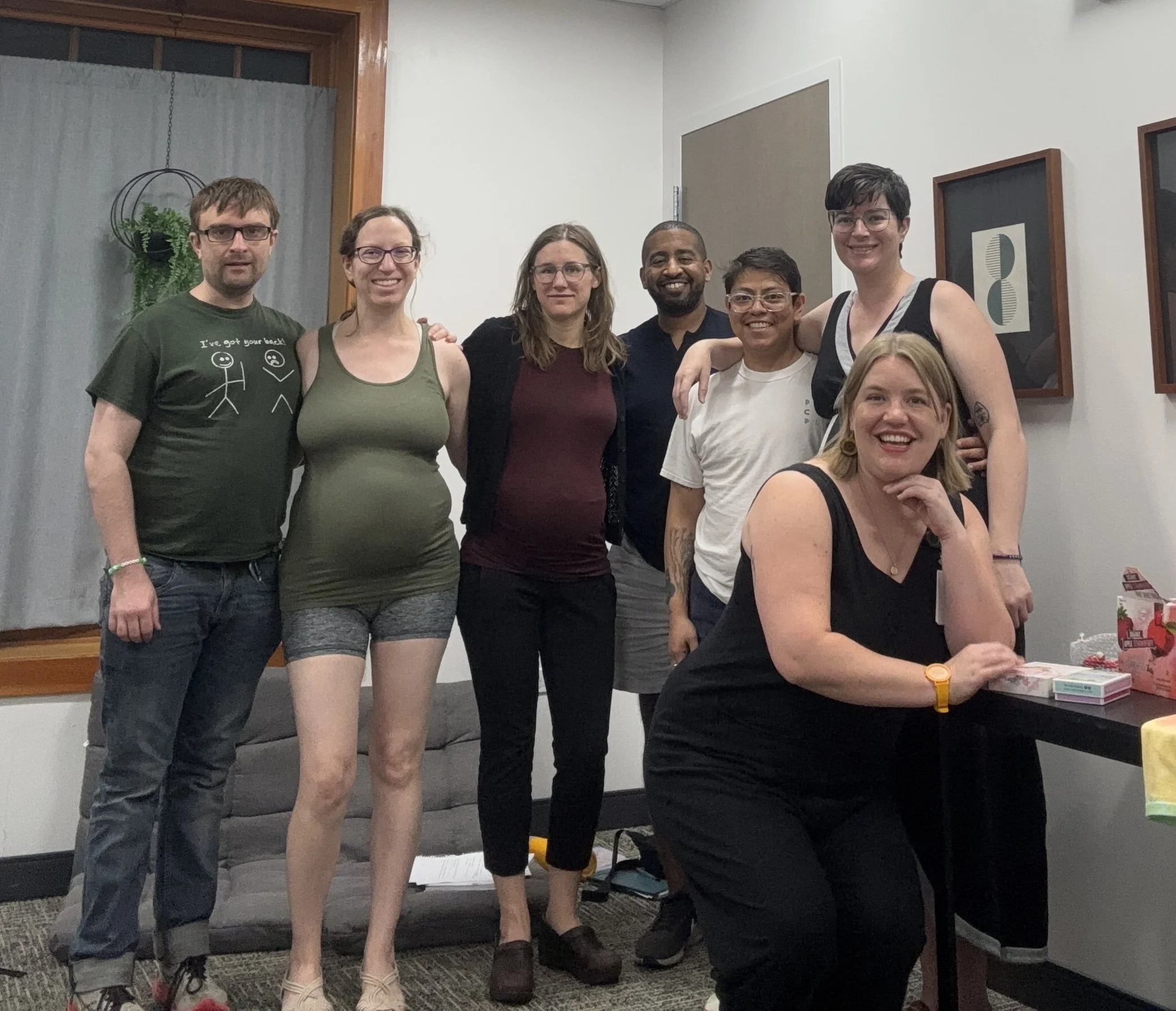 Group of five diverse pregnant couples posing together inside a room with white walls, framed artwork, and a gray couch, smiling at the camera.