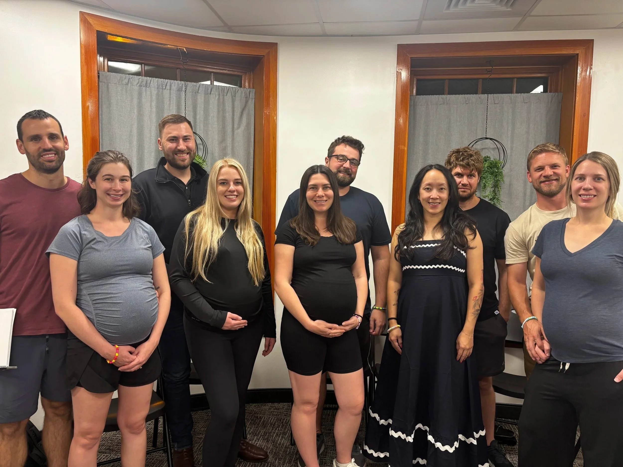 Group of 11 diverse pregnant couples indoors, some women are pregnant, standing in front of windows with wooden frames and gray curtains, smiling at the camera.