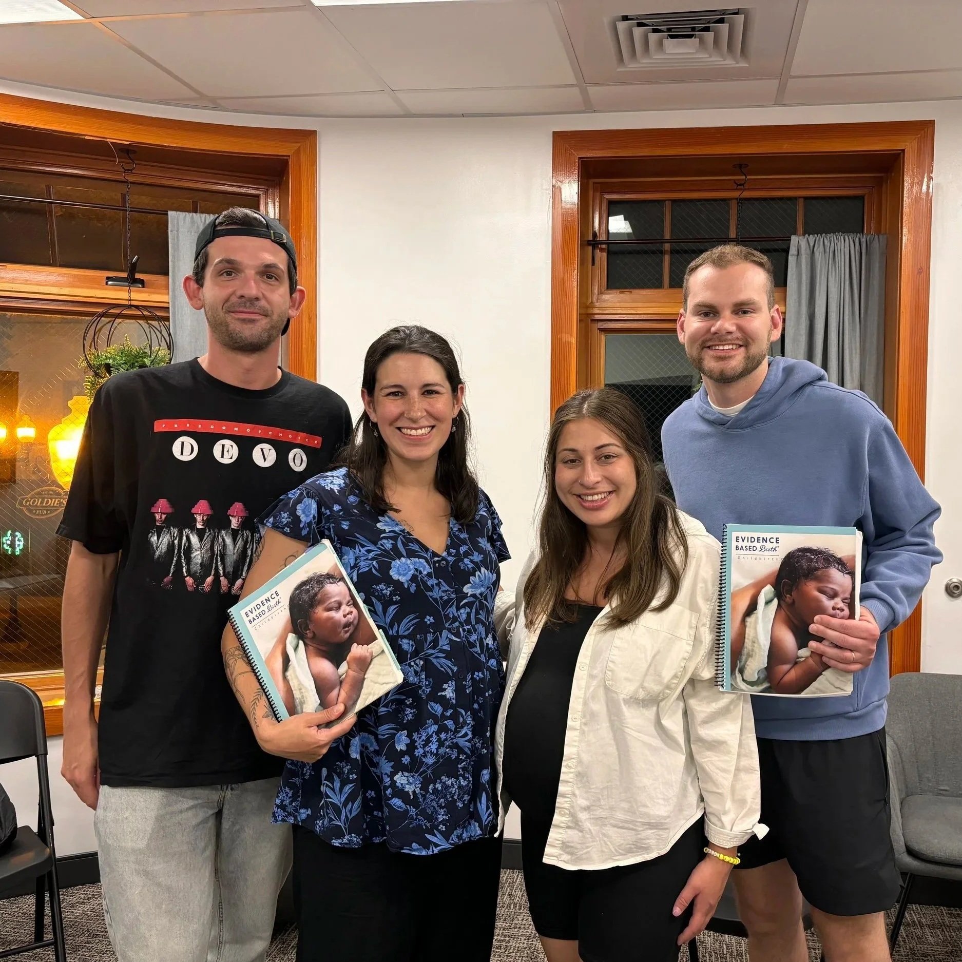 Two pregnant couples standing together indoors, two holding books with images of a sleeping baby on the covers, smiling at the camera.
