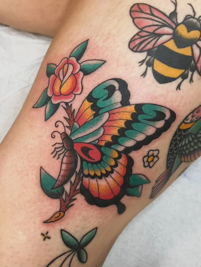Tattoo of a colorful butterfly with pink, orange, and blue wings, perched on a red and pink flower with green leaves. There is a bee tattoo above the butterfly and part of a bird tattoo visible to the right.