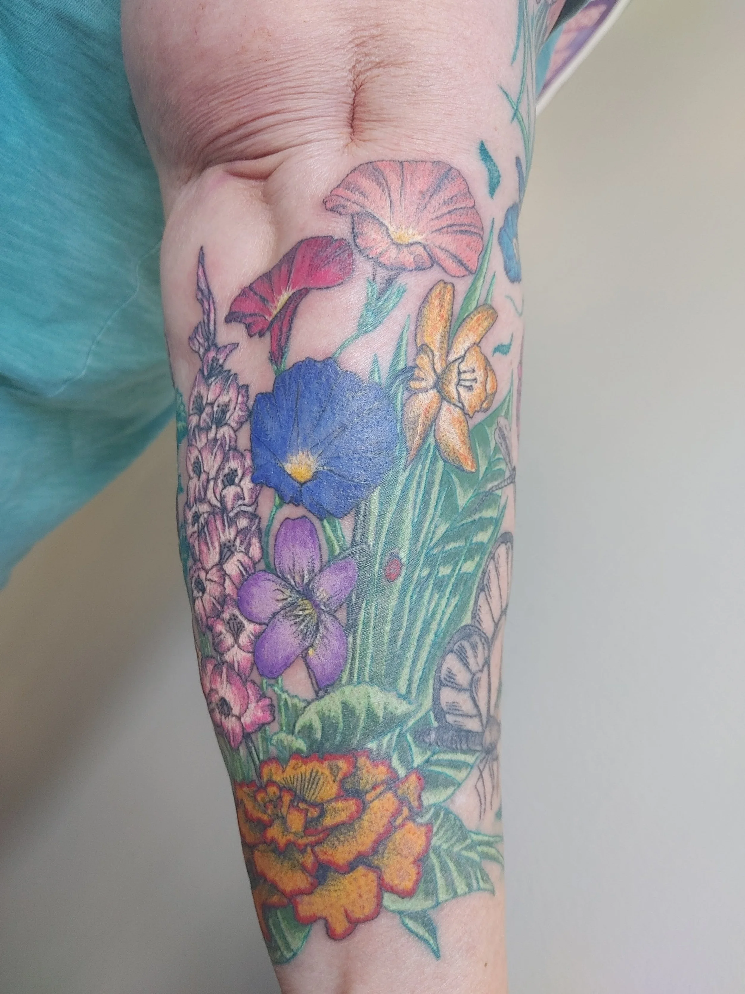 Close-up of a person's arm with a colorful tattoo of various flowers including a large purple flower, orange and yellow flowers, and green stems and leaves.