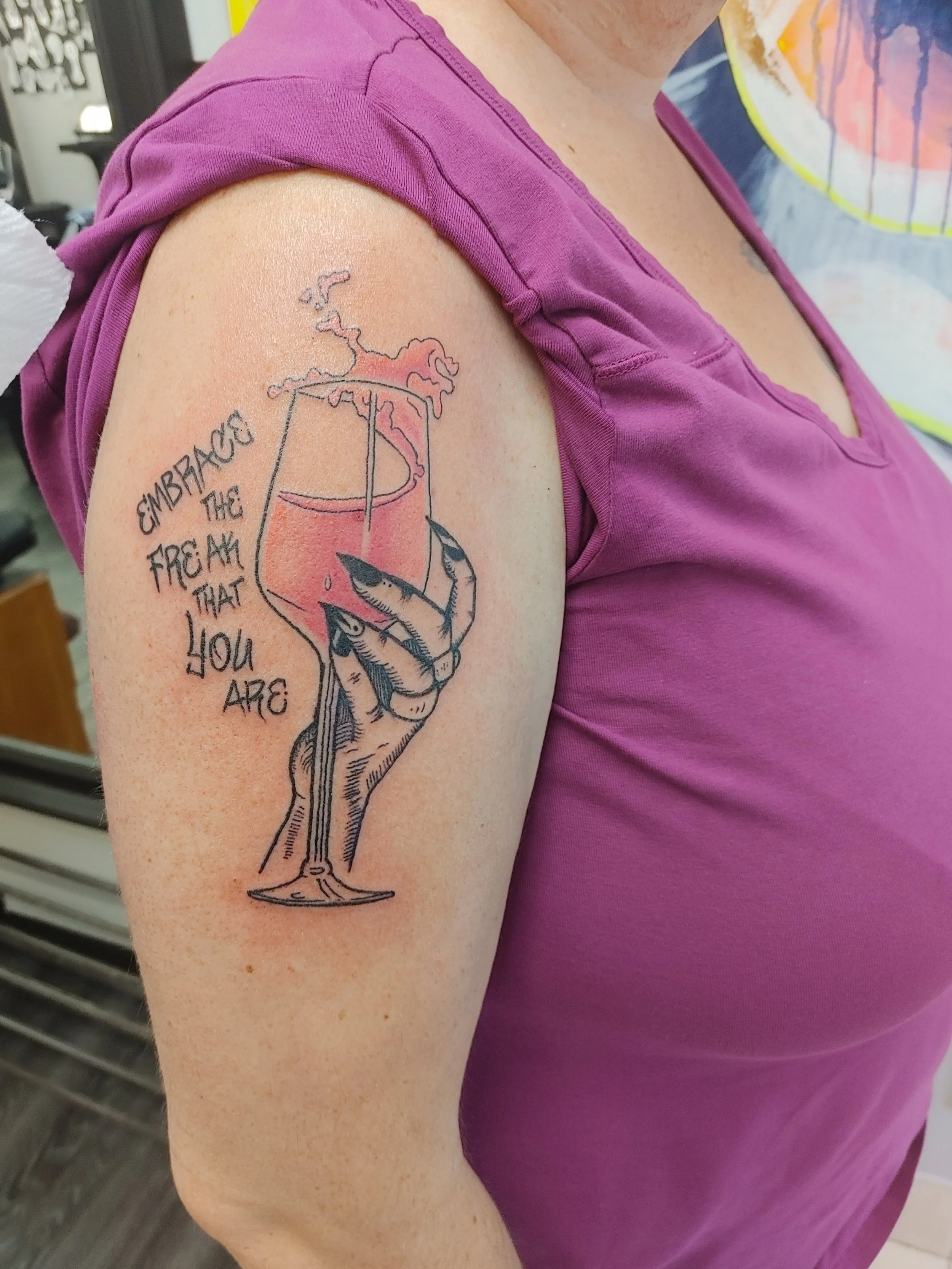 Tattoo of a glass of wine with a butterfly inside and the words "Gambace the freak that you are" on the upper arm, worn with a purple short sleeve shirt.