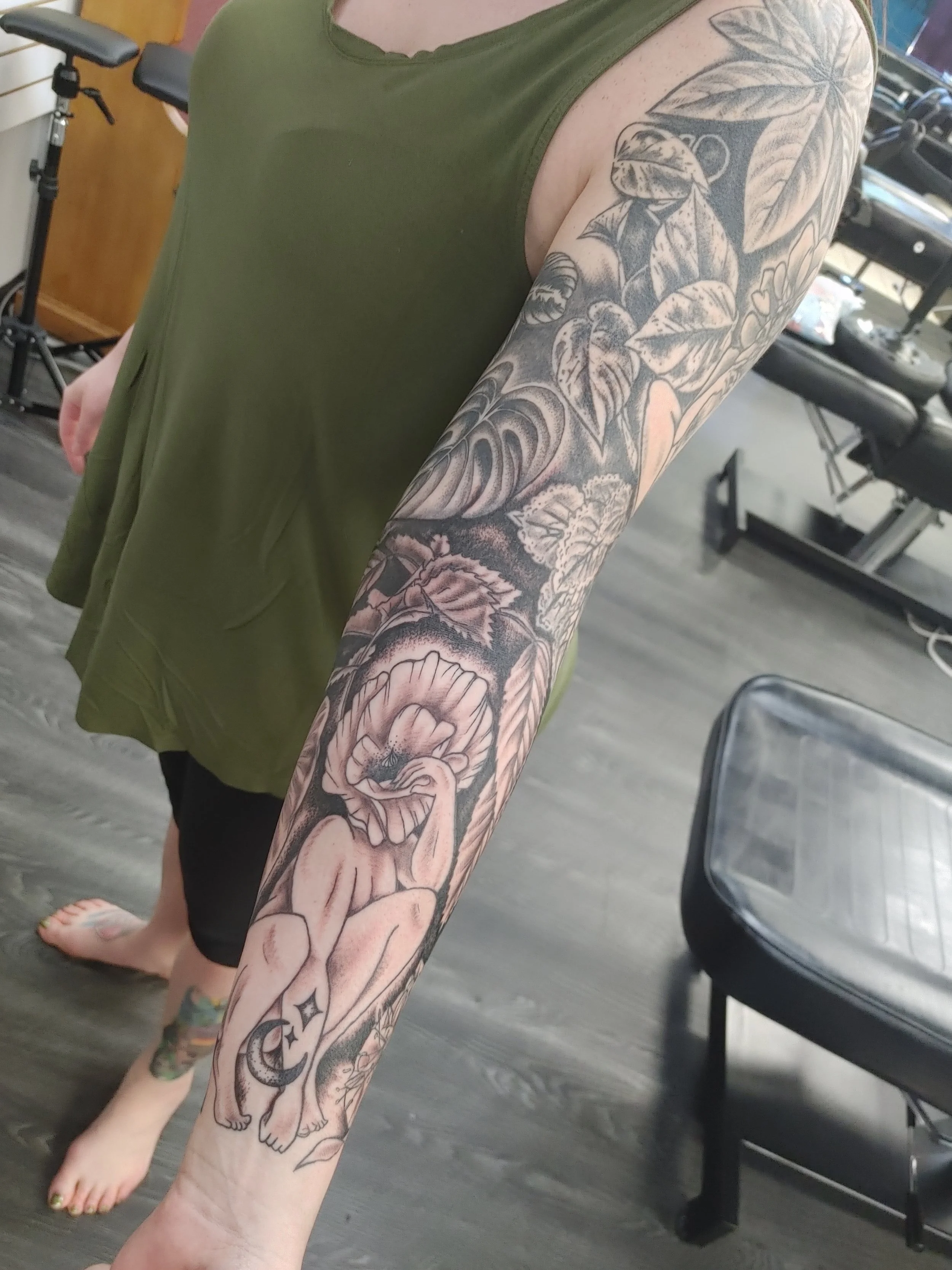A person's arm and lower leg with detailed black and gray tattoos of flowers, leaves, and a praying figure. They are standing in an indoor gym with workout equipment visible in the background.