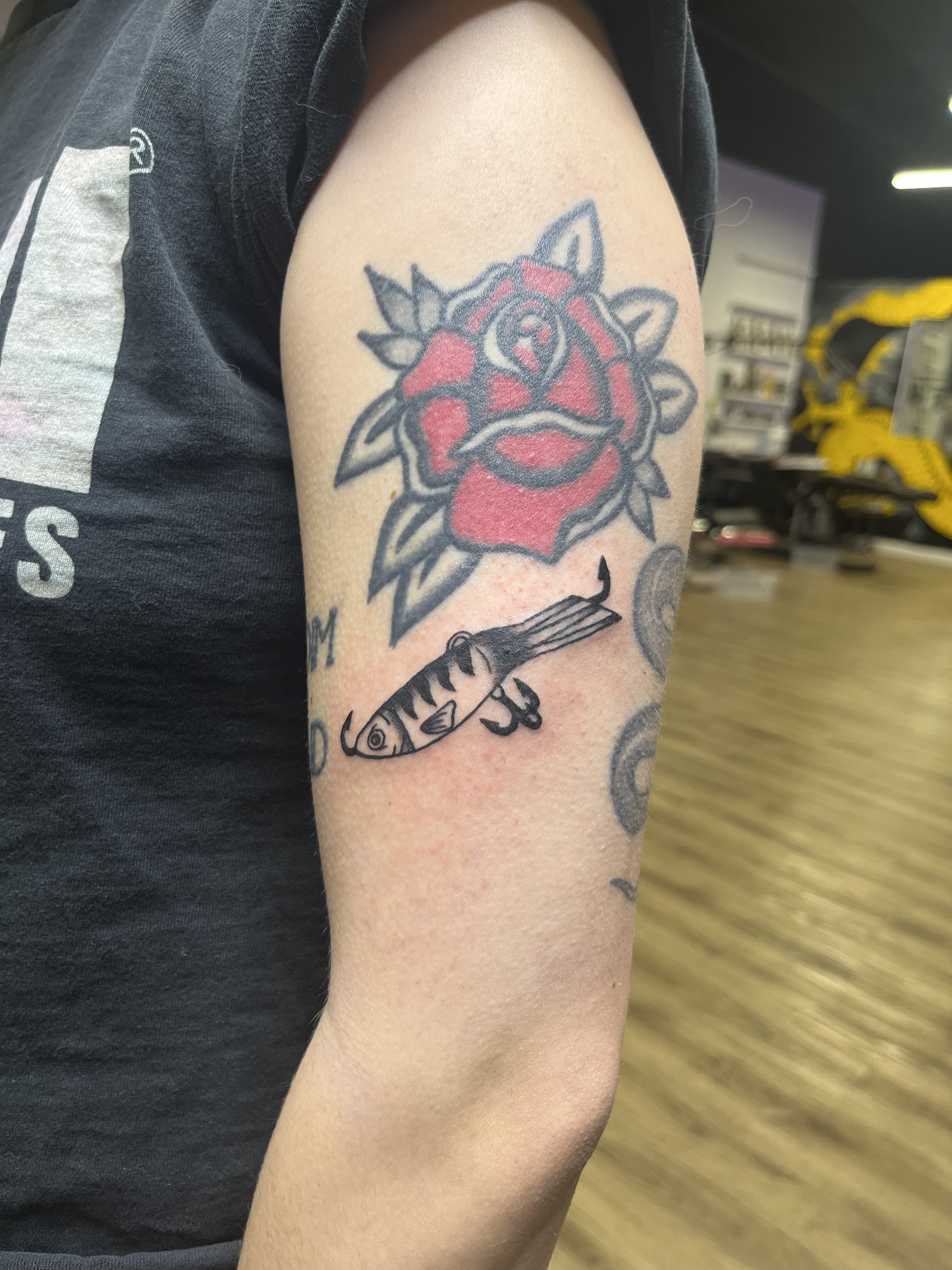 Tattoo of a red rose with leaves, a fish swimming below, and text that is partly visible.