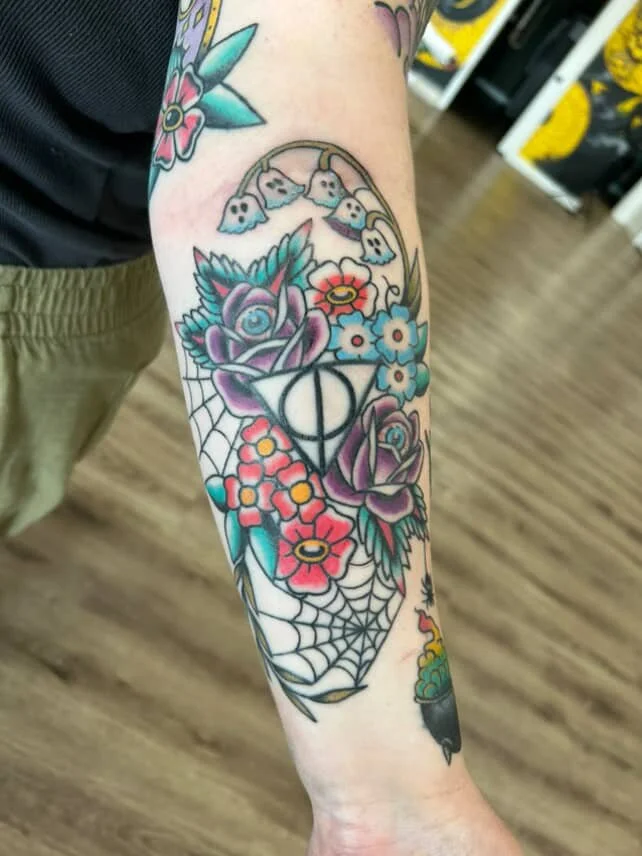 Colorful tattoo on forearm featuring roses, blue flowers, a spider web, a geometric symbol, and hanging ghost-like faces.