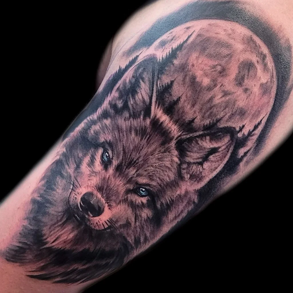 Tattoo of a fox with piercing blue eyes, moon in the background, and pine trees surrounding the fox on a person's arm.