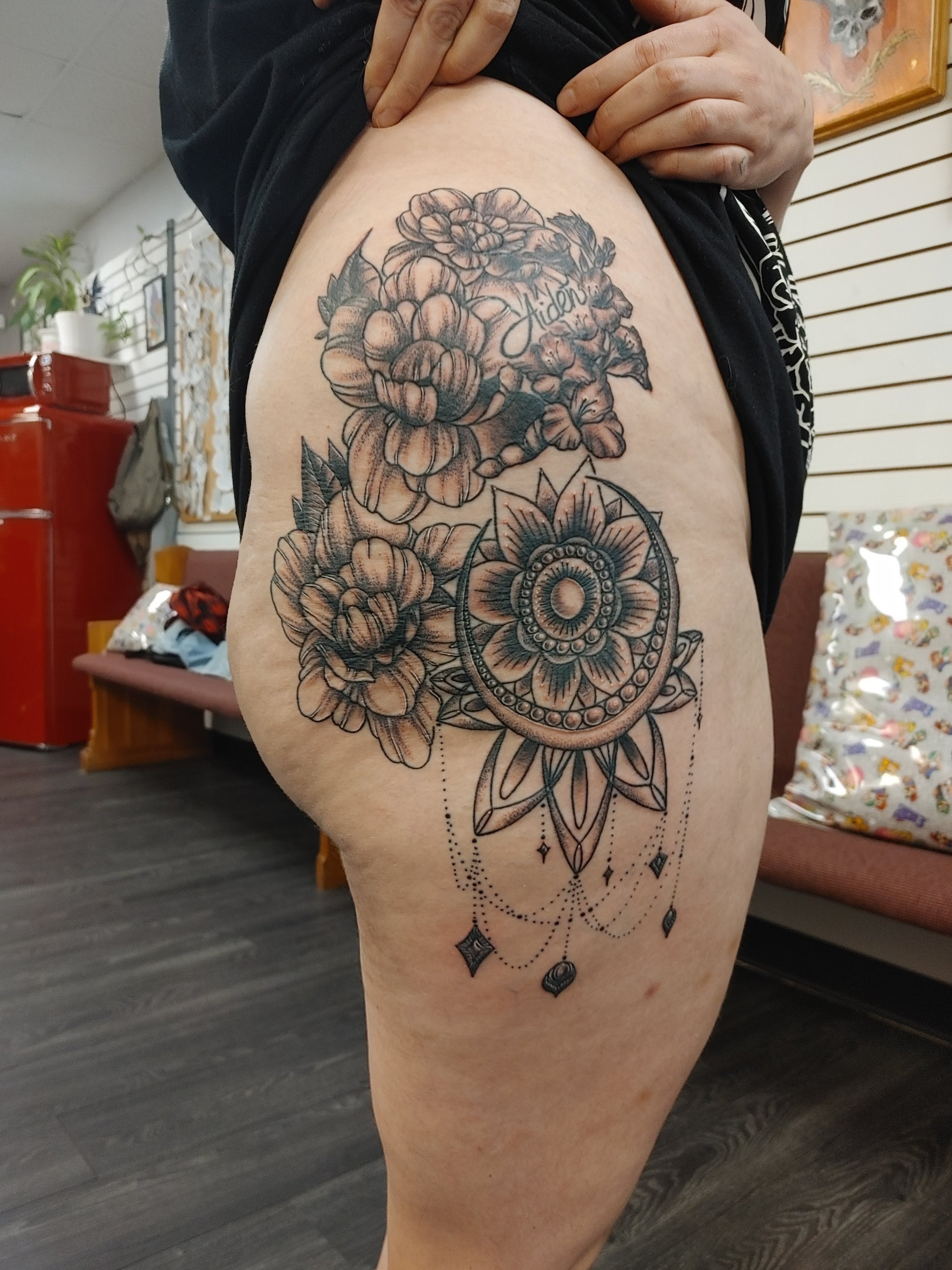 A person showing a thigh tattoo of a large floral design, featuring intricate flowers, leaves, and a decorative circular element with dangling diamond shapes and dotted lines.