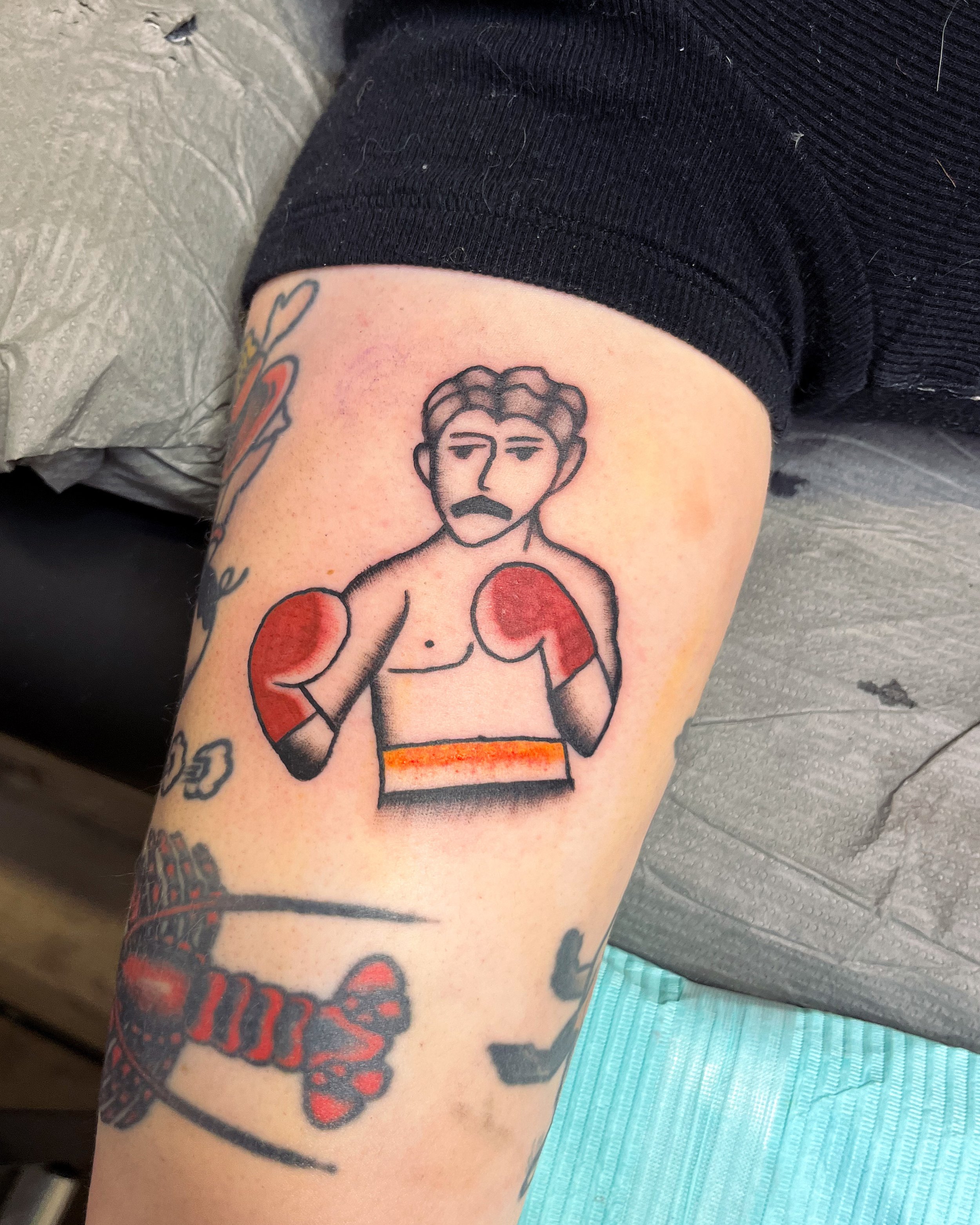 Tattoo of a boxing figure with blonde hair, a mustache, red gloves, and a determined expression on the person's arm.