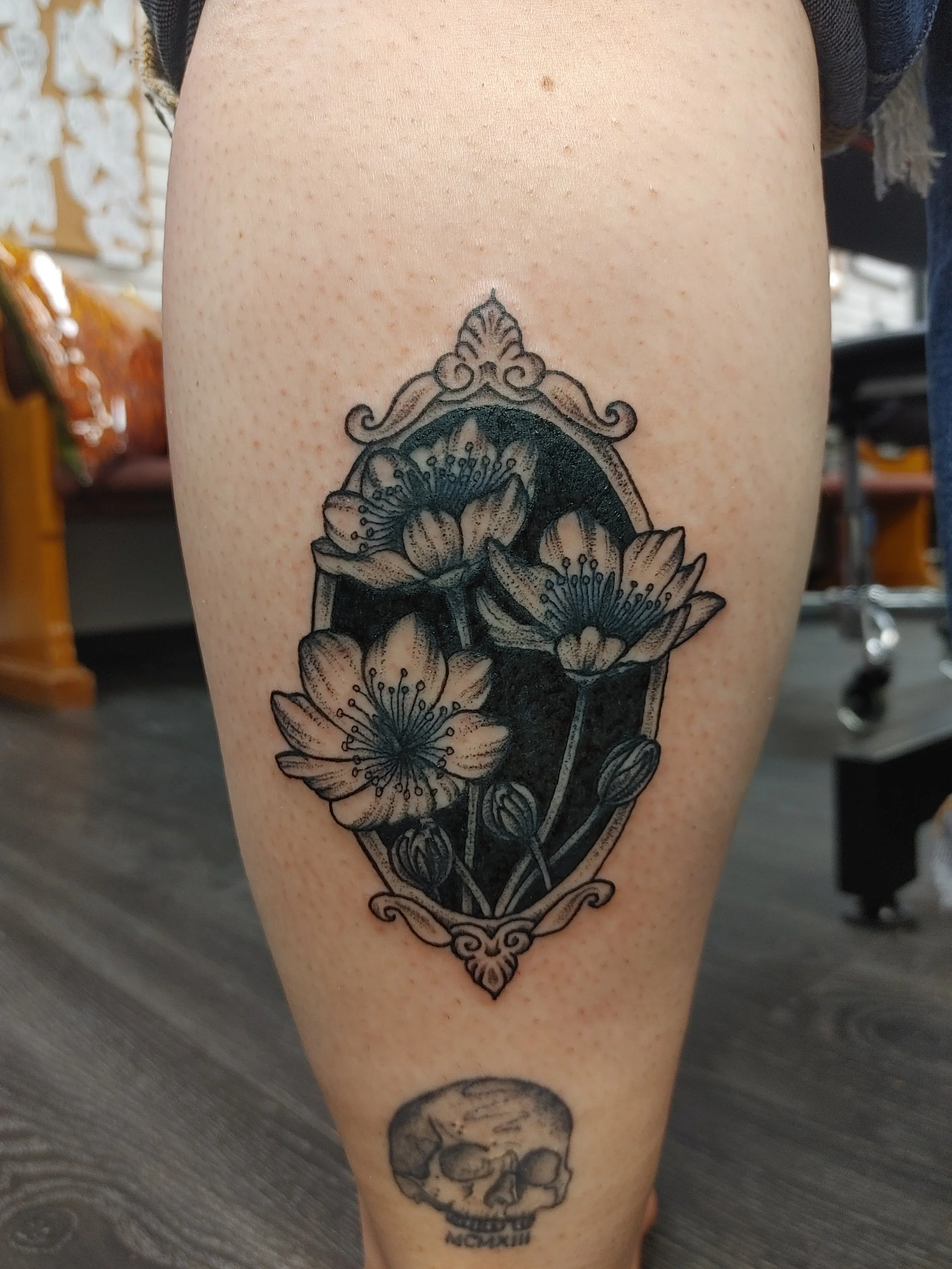 Tattoo on a person's leg featuring a floral design with flowers and leaves inside an ornate frame, and a skull below with the word 'MOM' underneath.