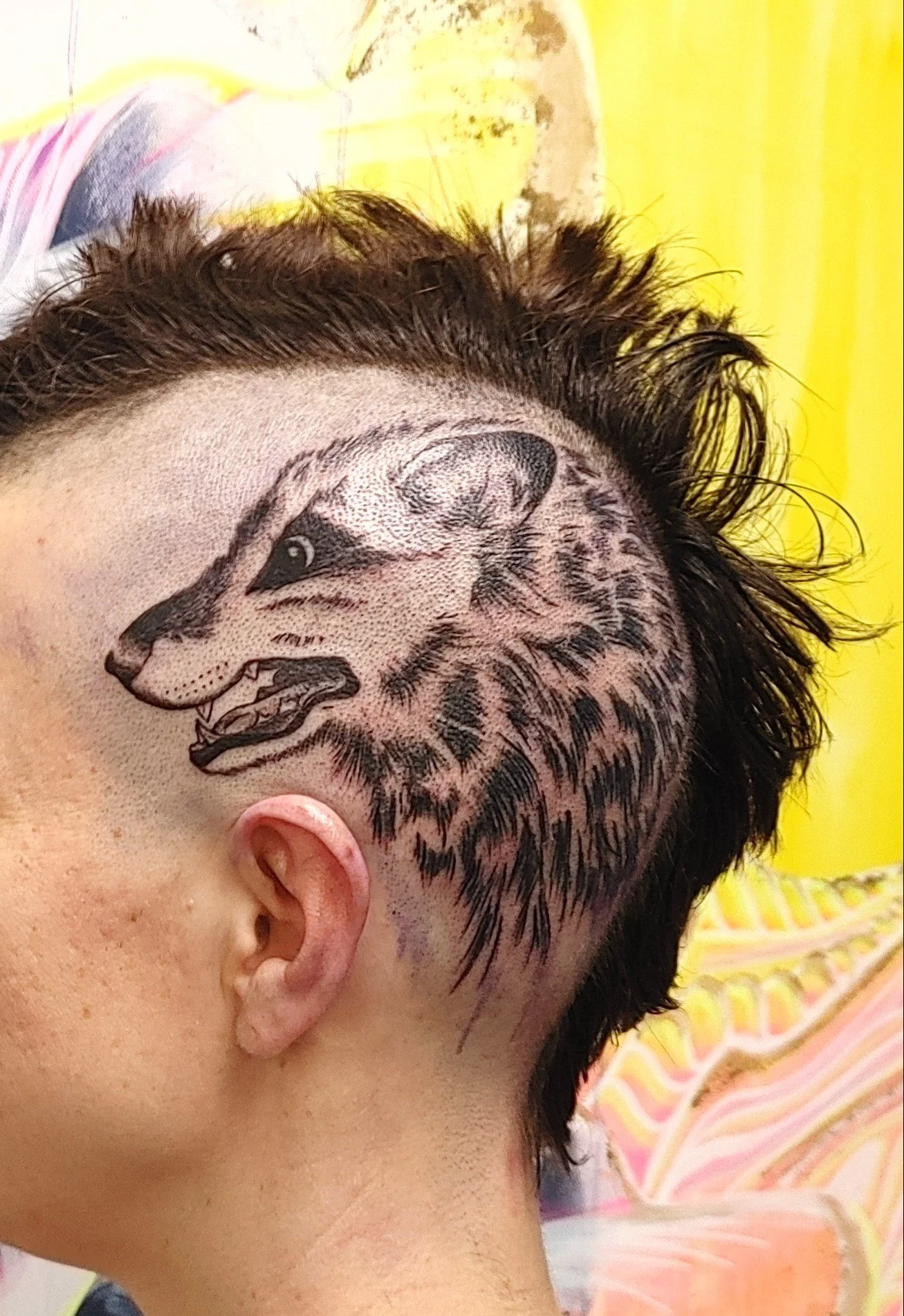 A person's face with a detailed tattoo of a wolf's head on the side of their face, their hair is dark and curly, and they are lying on a colorful pillow with yellow and pink patterns.