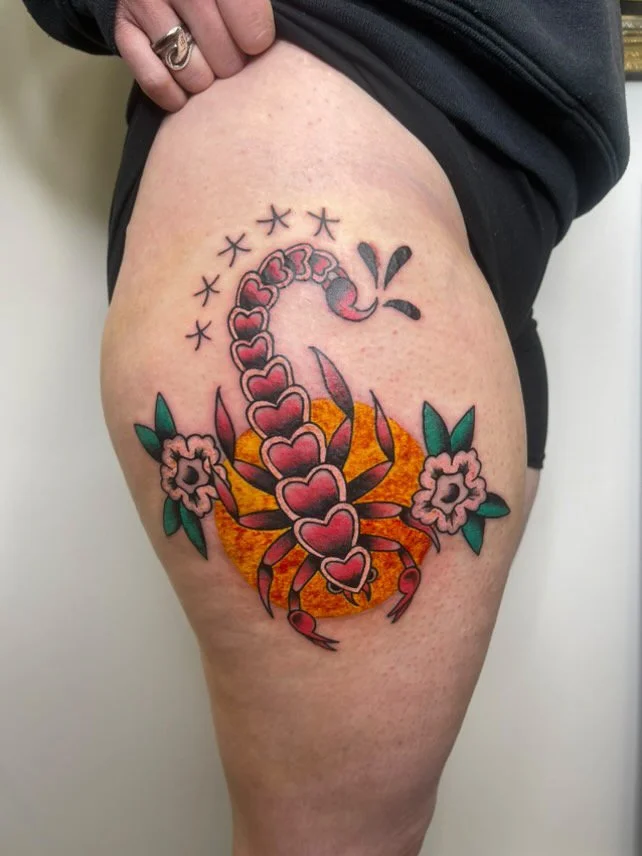 Scorpion tattoo with hearts, flowers, and a sun or fiery background on a person's thigh.