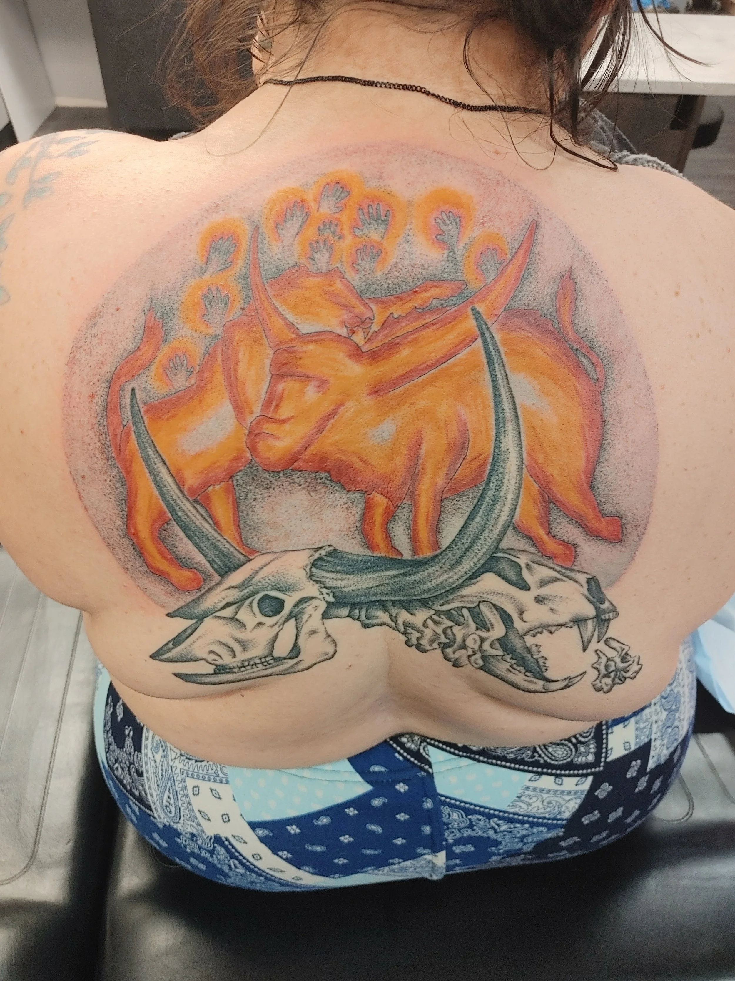 Colorful tattoo on shoulder and upper arm featuring a dragon with a crown. The tattoo incorporates various colors including orange, gray, and black, with additional elements such as small hand motifs and leaf-like designs.