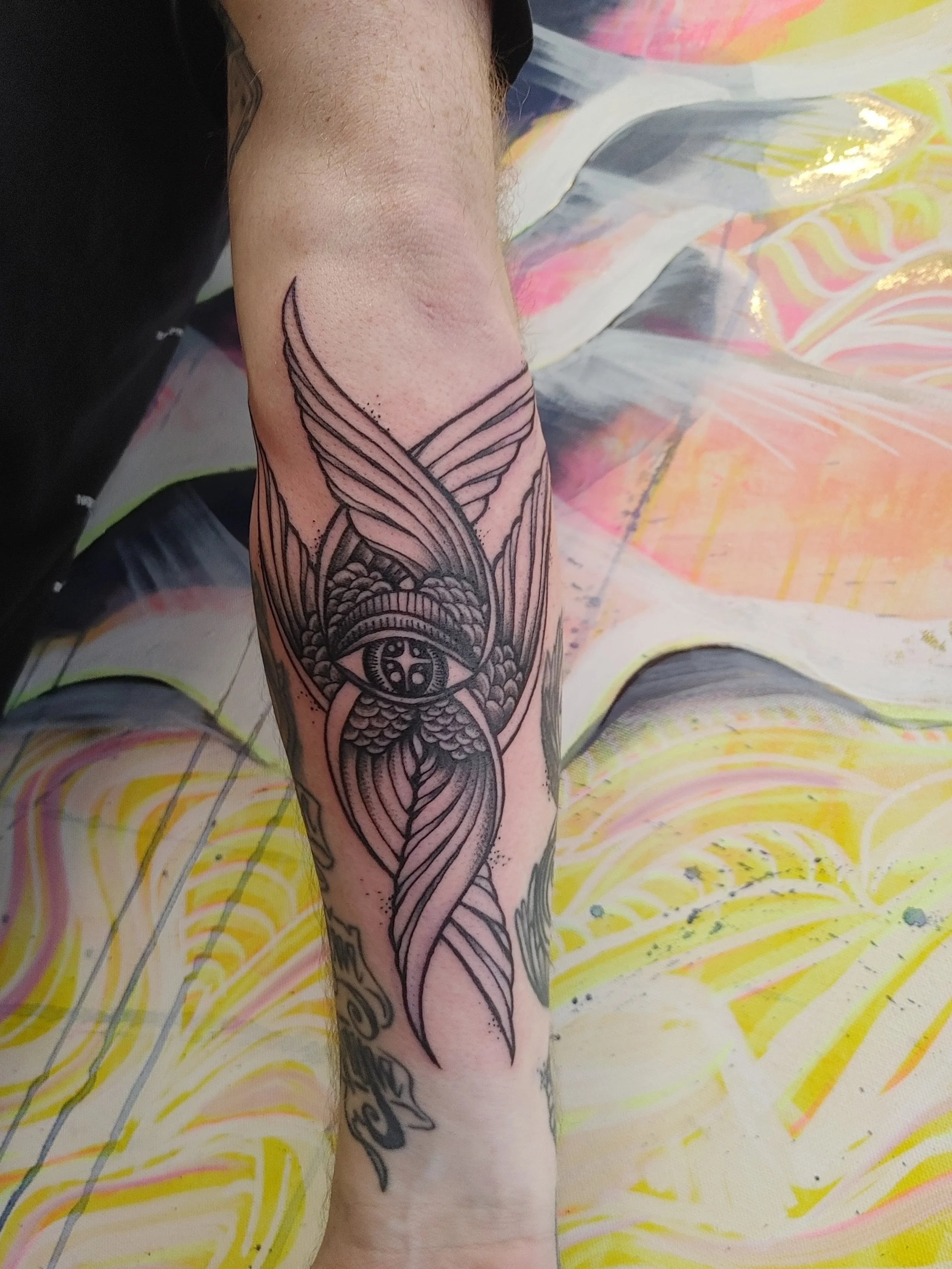A person's arm with a detailed black ink tattoo of a winged insect, possibly a moth or butterfly, with intricate patterns and a face within the design, resting on a colorful, abstract painted surface.