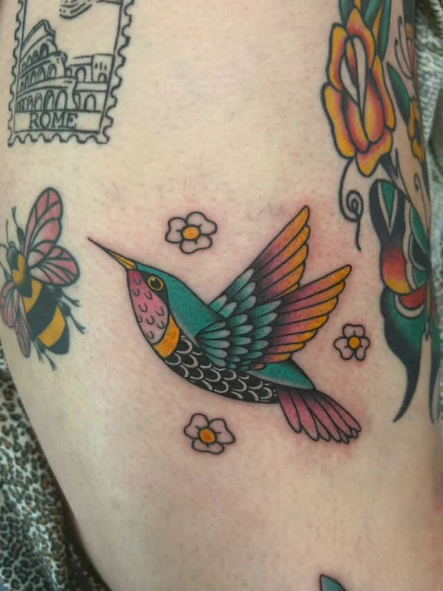 Colorful tattoo of a hummingbird surrounded by small flowers, a butterfly, and part of a tattoo depicting Rome.