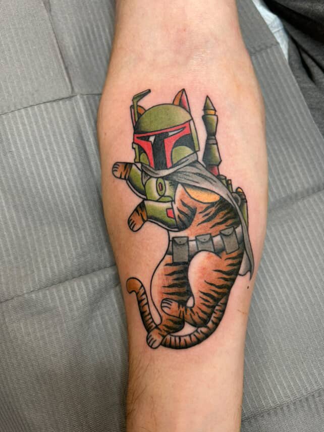 Tattoo of a character with a helmet and armor holding a cat with a gray bandana, the cat is orange with black stripes, and the character appears to be from the Star Wars universe, resembling Boba Fett.