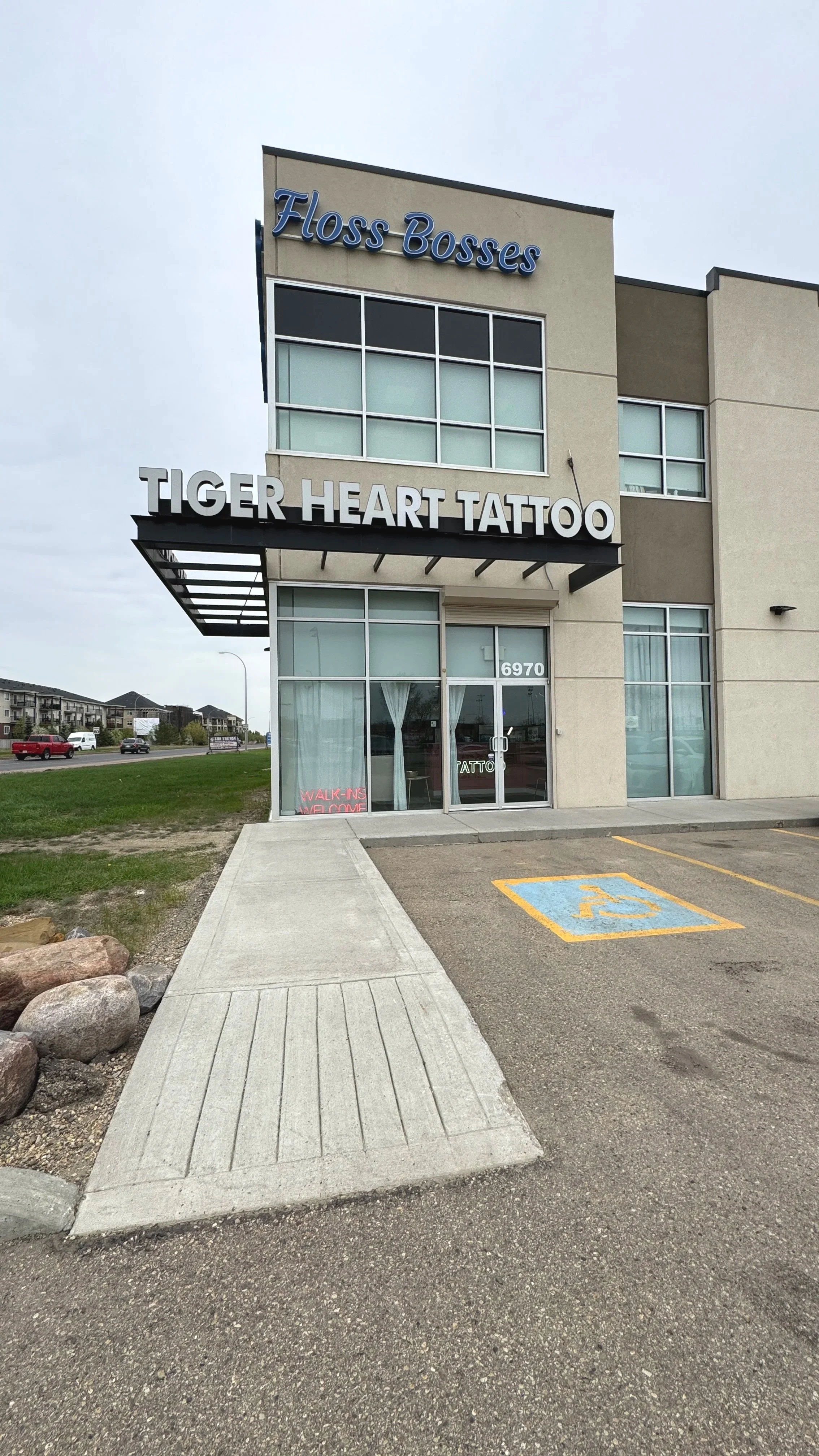 Exterior of a commercial building with signs reading "Floss Bosses" and "Tiger Heart Tattoo." There is a parking lot with a designated handicap parking spot and a sidewalk leading to the entrance.