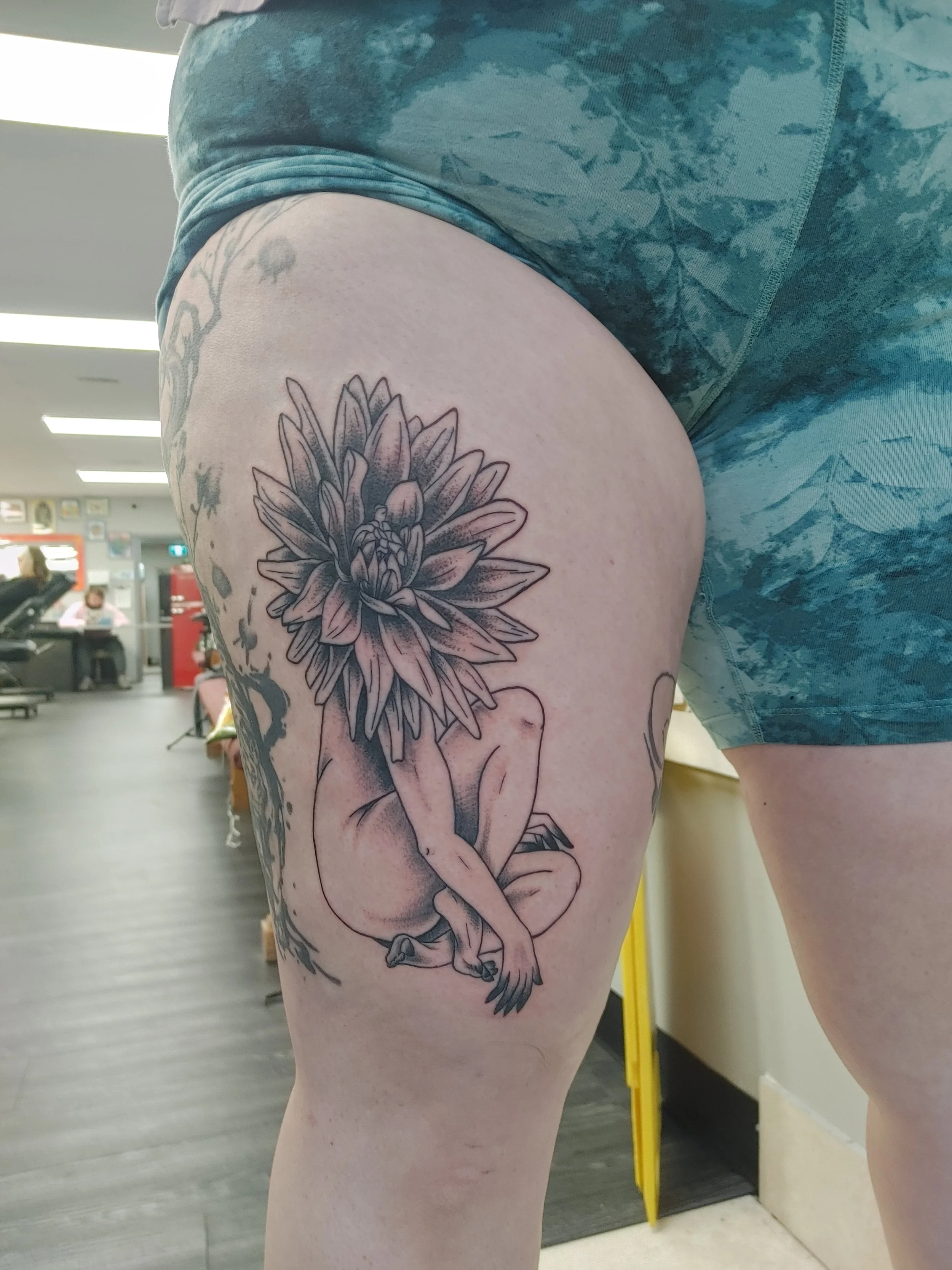 A woman with a large floral tattoo on her thigh, seated in an indoor space that looks like a gym or waiting area.