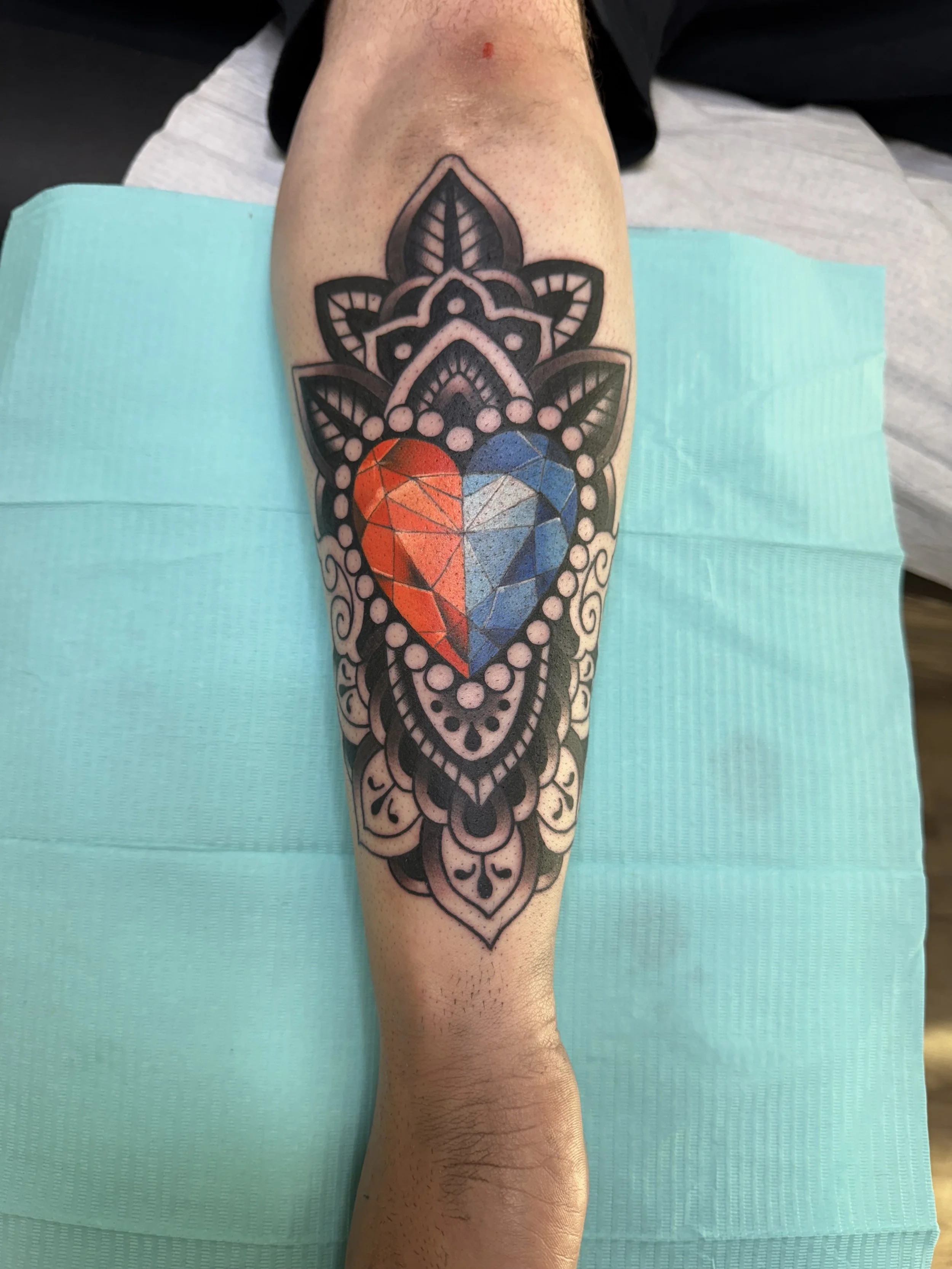 Tattoo of a colorful geometric heart inside a black ornate design on a person's leg.