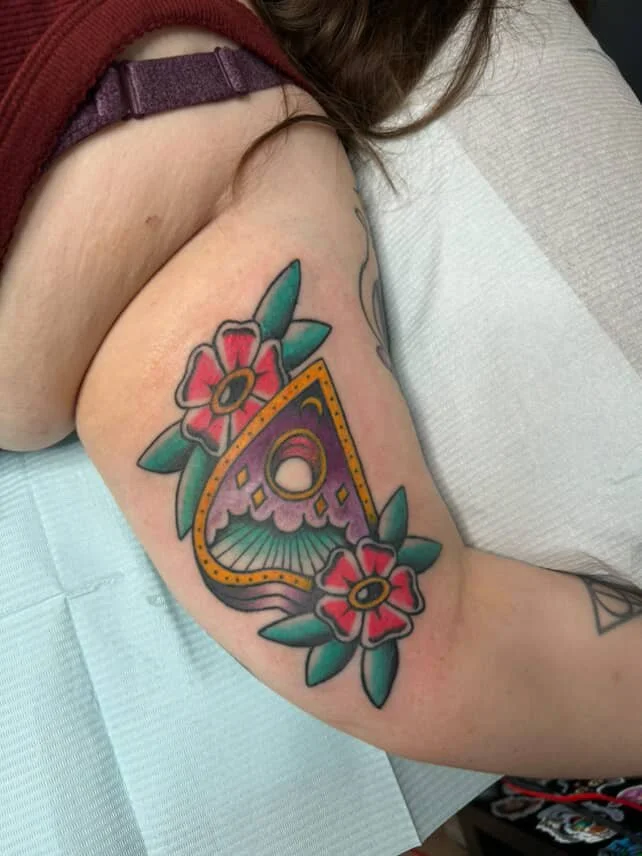 Colorful tattoo of a stylized eye within a geometric shape, surrounded by pink flowers with green leaves on a person's upper arm.