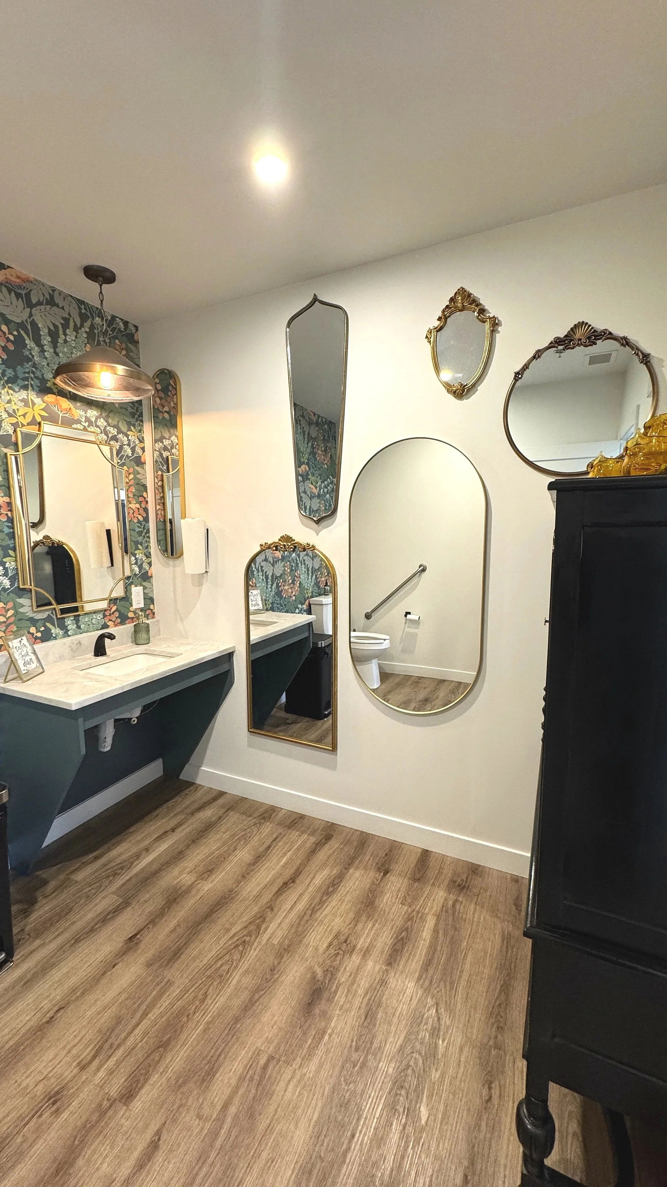 Decorative bathroom with vanity sinks, multiple mirrors on the wall, wallpaper with floral pattern, wood flooring, and a toilet visible through an arched opening.