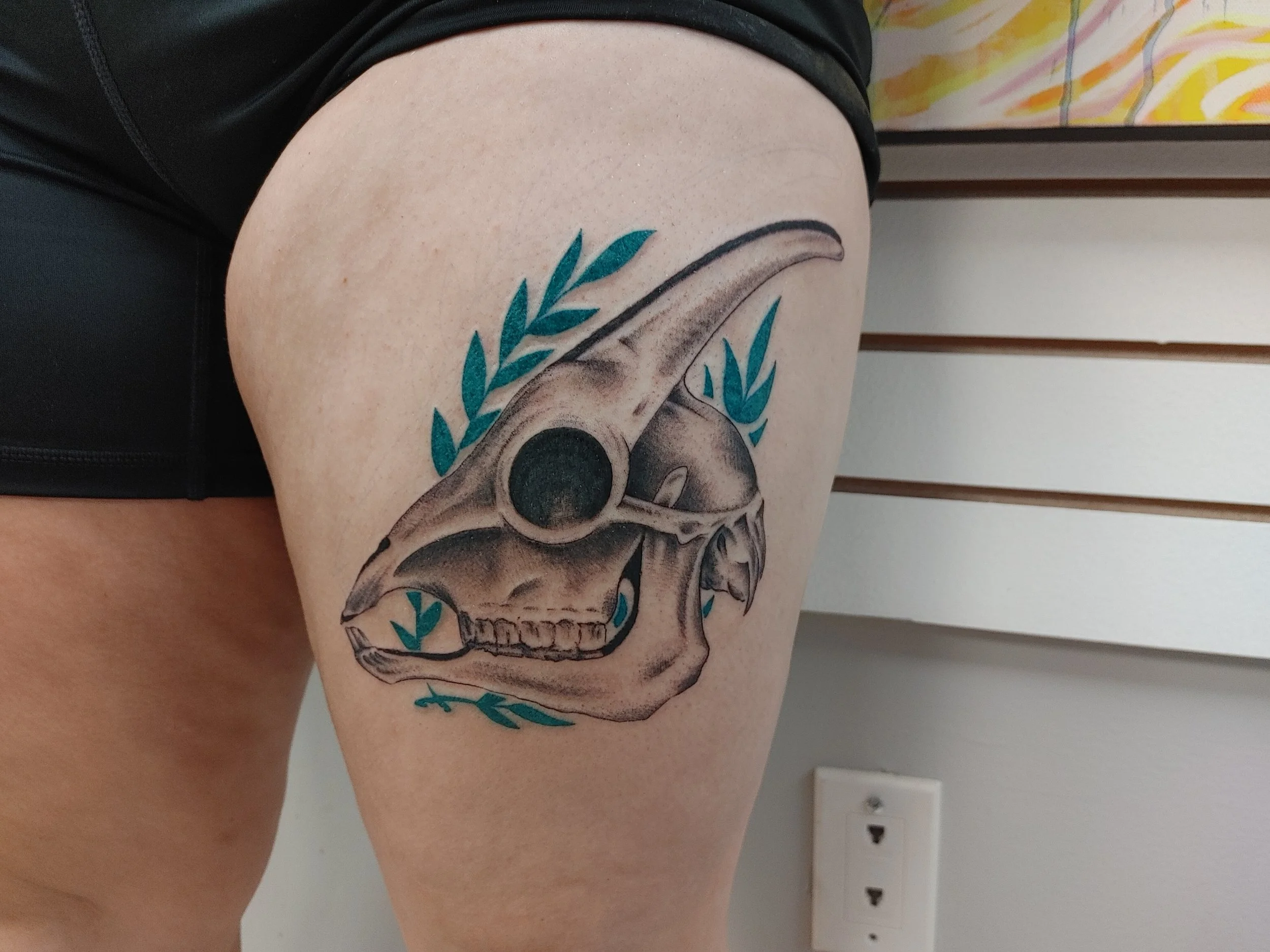 Tattoo of a skull with leafy accents on a person's thigh.